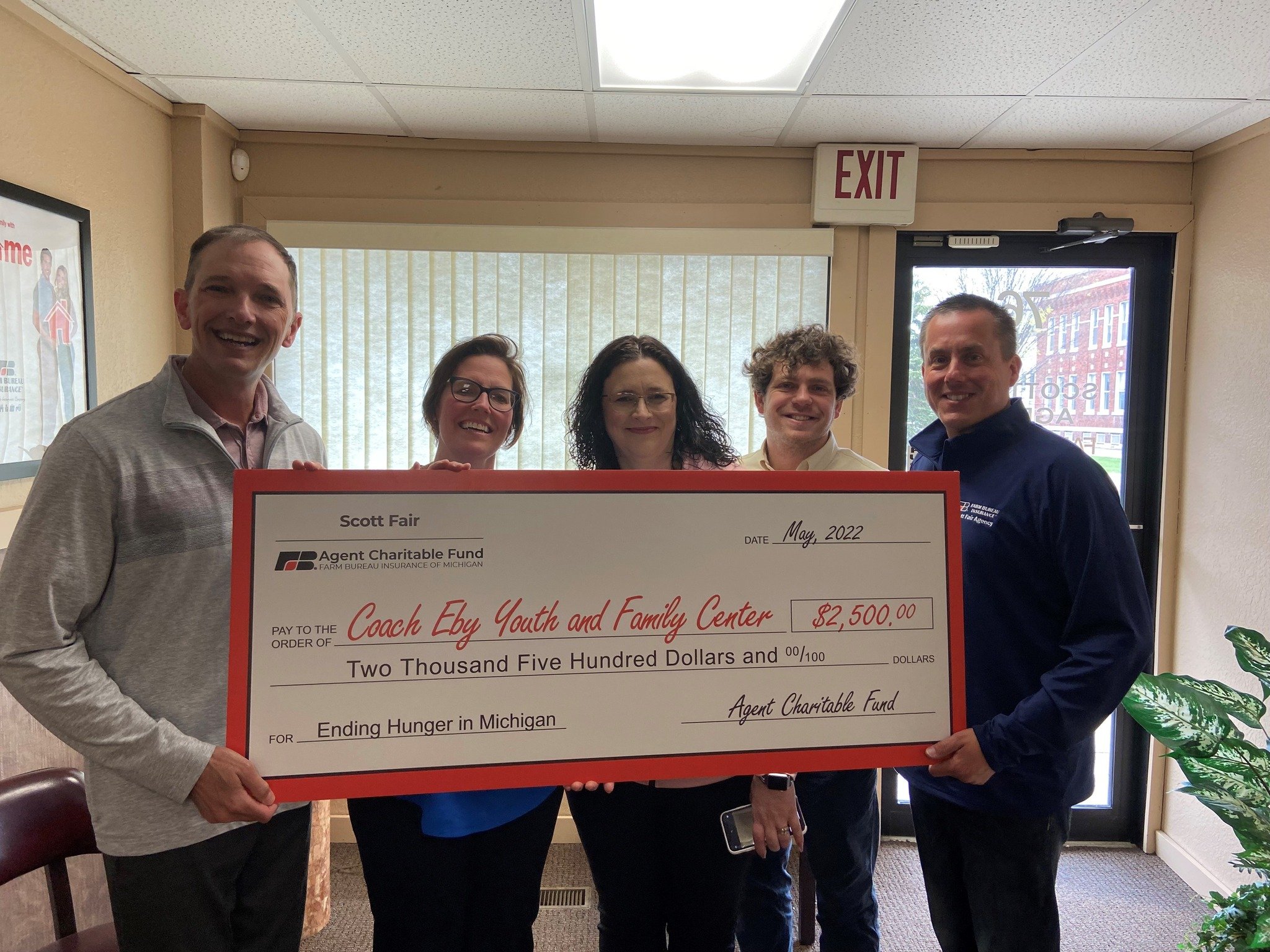 A group of five people holding a large check for $2,500, standing in a room with windows and an exit sign overhead.
