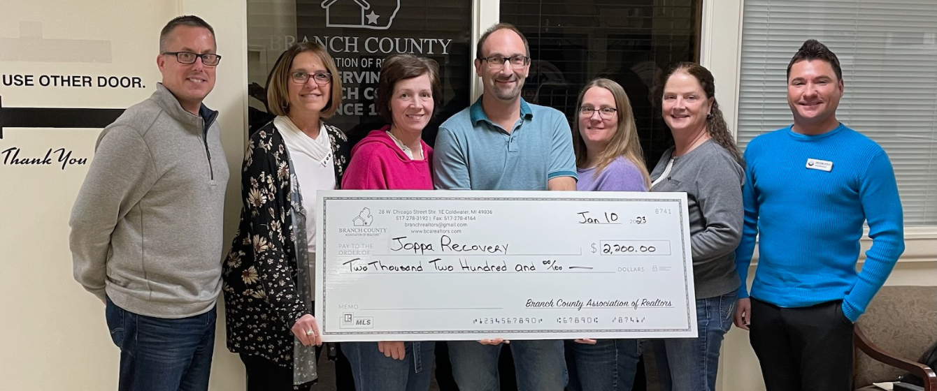 Group of seven people standing side by side in an office, holding a large check made out to Joppa Recovery for $2,200, with the date January 10, 2023. The check is from the Branch County Association of Realtors.