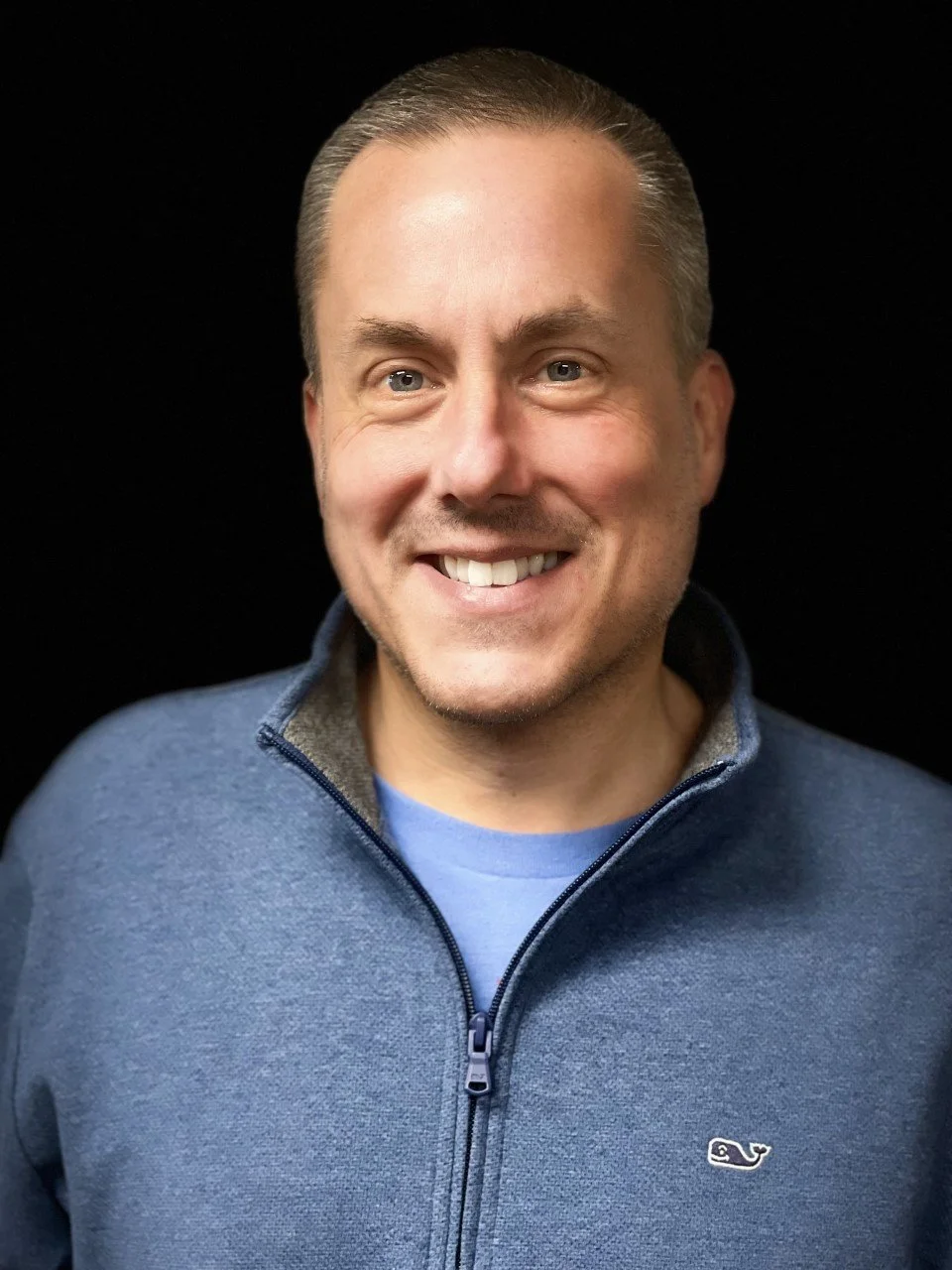 A man with short dark hair and a beard, wearing a navy blue polo shirt with the Farm Bureau Insurance logo and text that reads "Aird Garcia Insurance Group," standing in front of a light-colored brick wall.