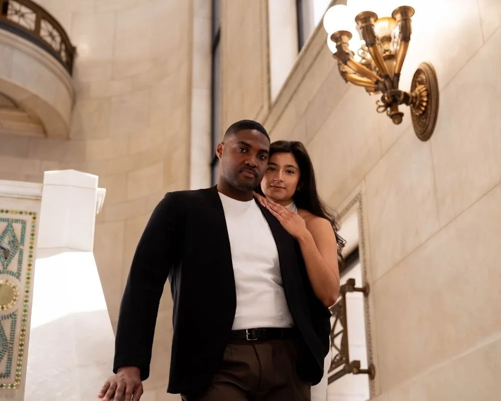 A young man and woman posing together inside a building with elegant, beige walls, and vintage light fixtures.