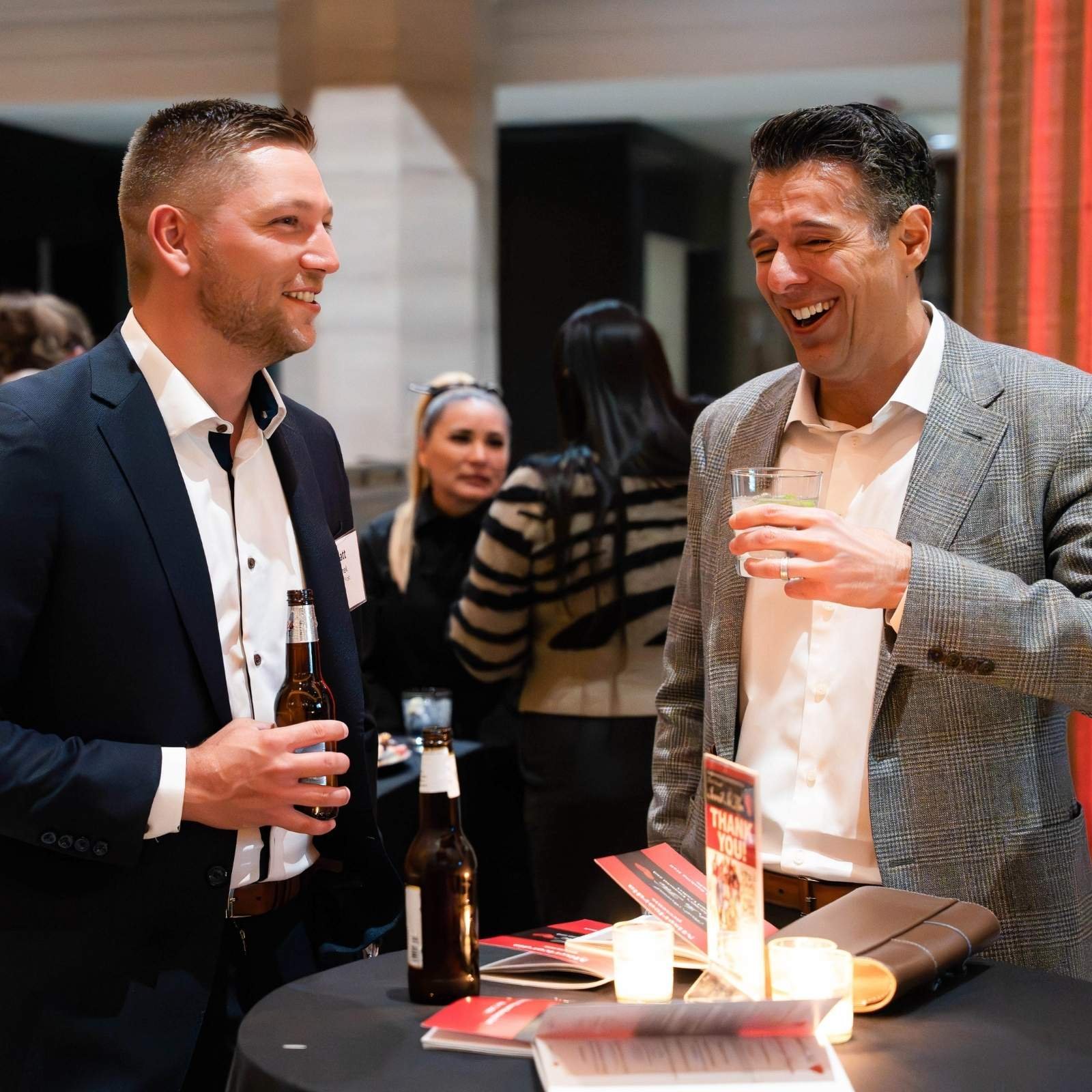 Two men in formal attire are engaged in conversation at a social event, holding drinks. One has short dark hair and a beard, while the other has short black hair and is smiling broadly.