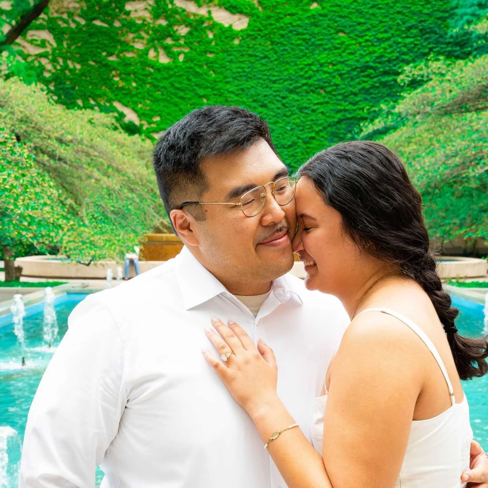 A couple sharing a close, affectionate moment outdoors near a fountain with lush green trees in the background.