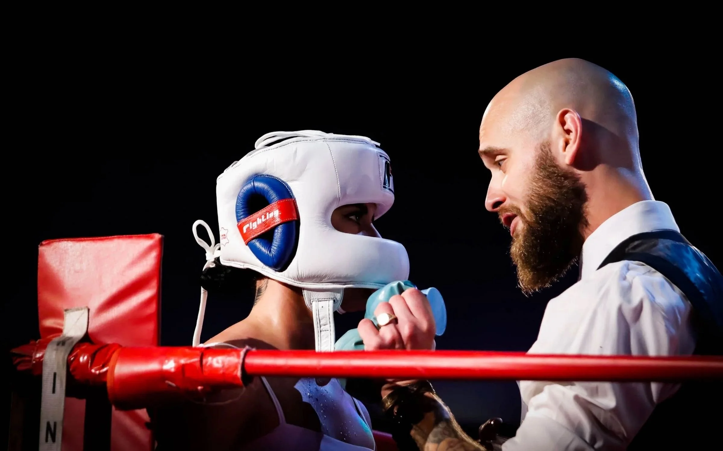 A boxer wearing protective headgear and gloves in a boxing ring faces a trainer or coach, who is whispering to the boxer.