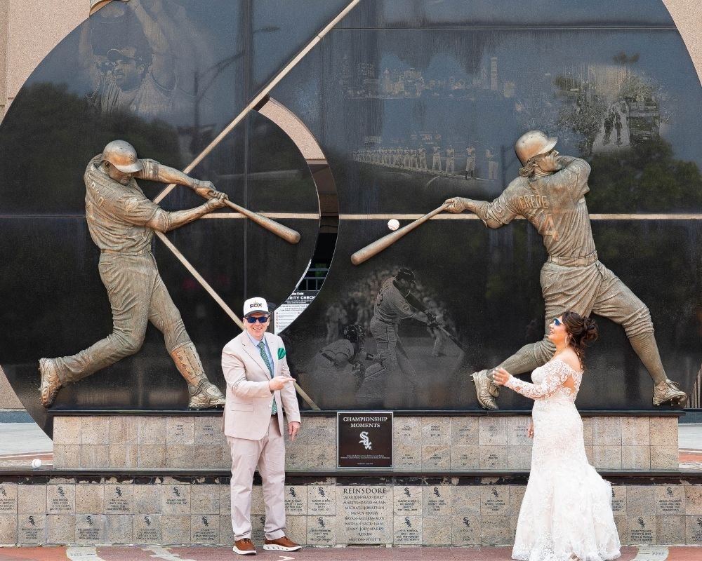 A woman in a white wedding dress and a man in a suit are posing in front of a large black baseball sculpture with bronze figures of baseball players. The sculpture features two players in a dynamic batting stance, and the couple appears to be holding baseball bats, fitting the theme.