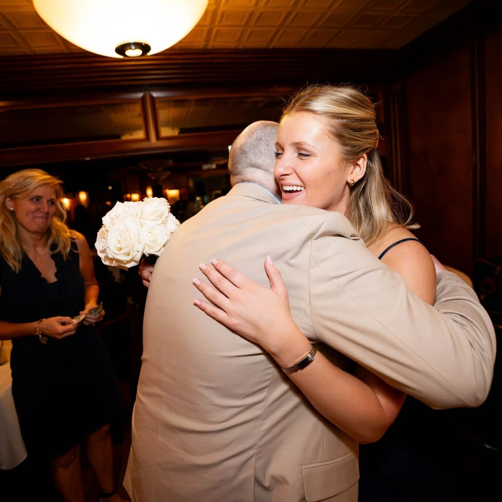 A woman with blonde hair smiling and hugging a man in a suit, holding a bouquet of white roses, in a warmly lit room with wood-paneled walls. A woman with blonde hair and a black dress is in the background.