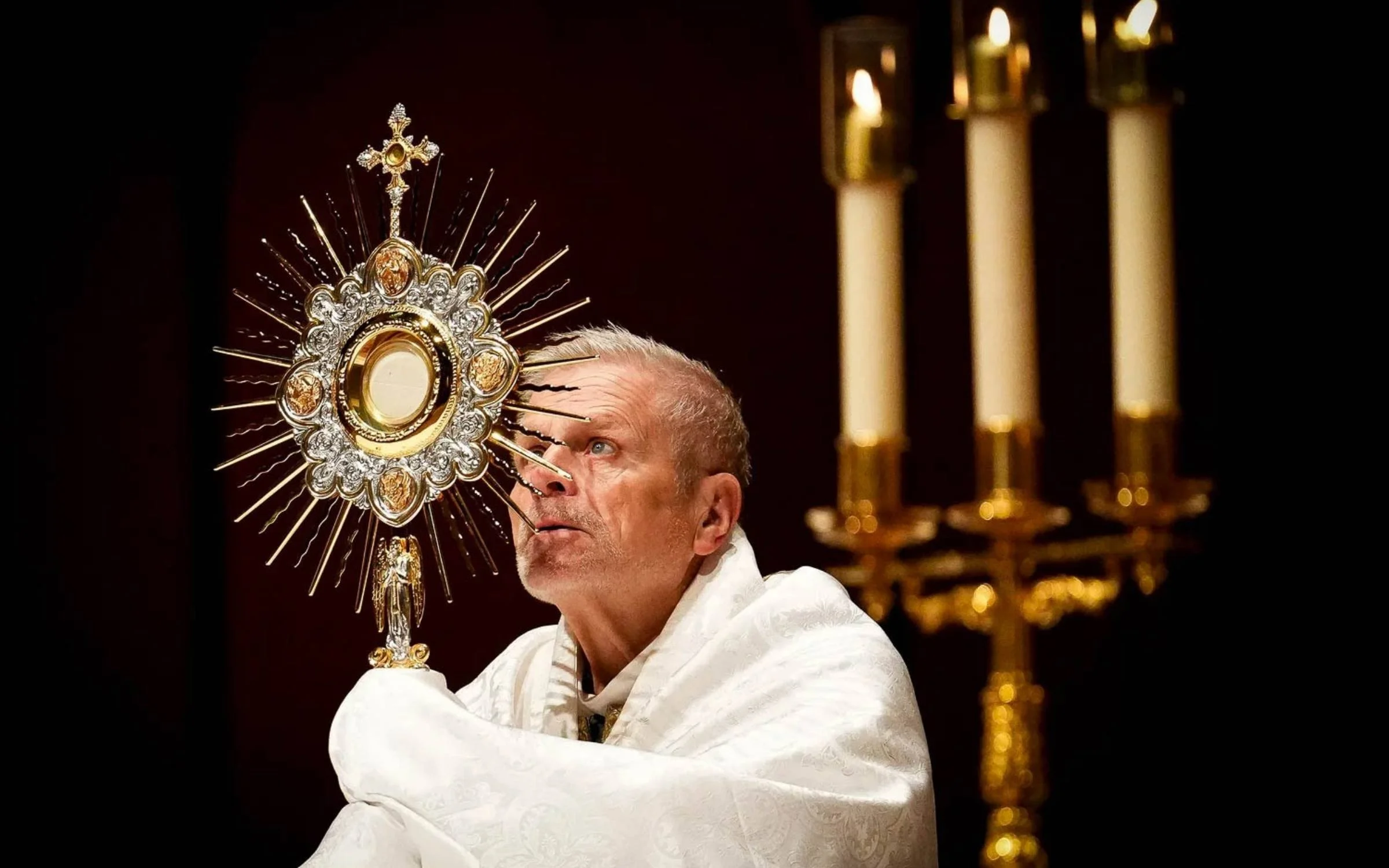 A clergy member holding a golden monstrance during a religious ceremony, with lit candles in the background.