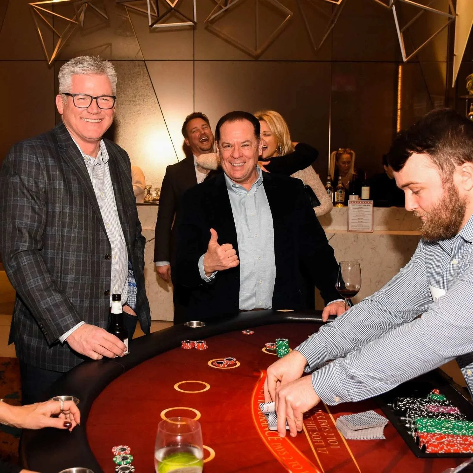 A group of people at a casino table playing poker, with some smiling and others focused on their cards, in an upscale setting with a dark background and geometric decorations.