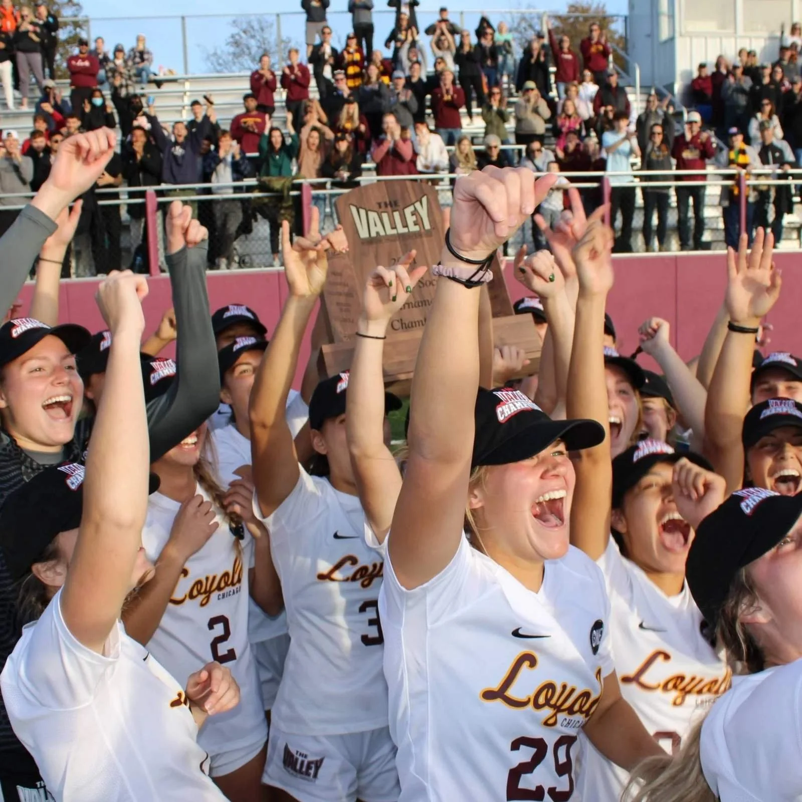 A group of female athletes wearing baseball uniforms celebrating victory after a game, holding a trophy, with spectators cheering in the background.