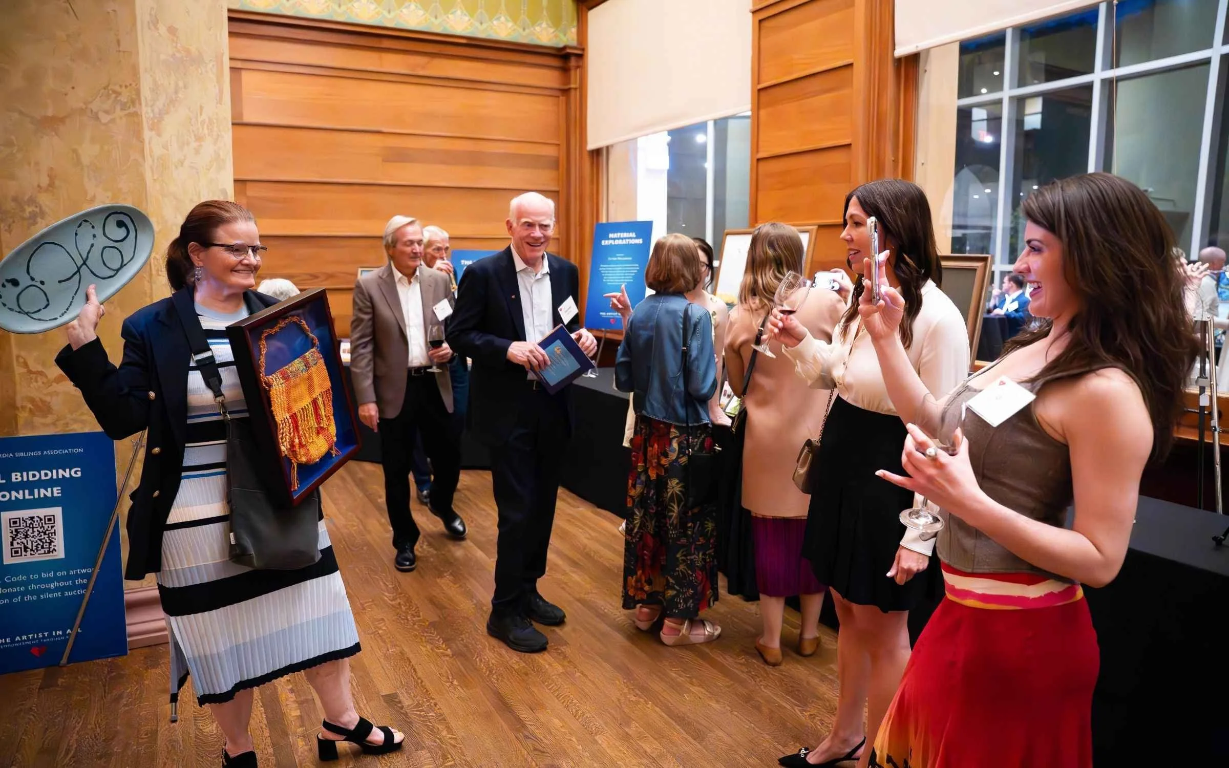 Group of people at an art auction or exhibition, some viewing artwork, smiling, talking, and taking photos in a room with wooden walls and large windows.