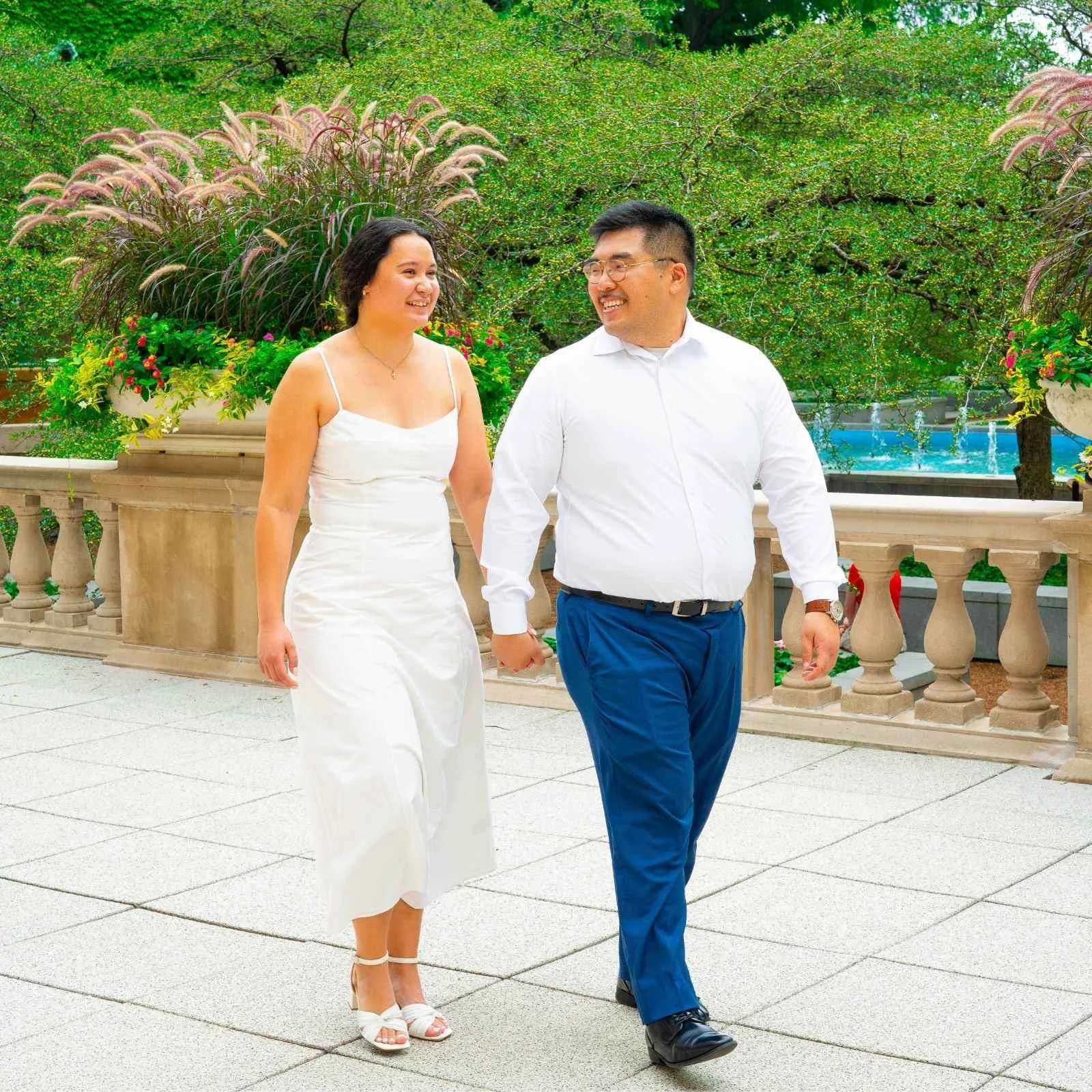 A couple holding hands and walking outdoors near a garden with green bushes and pink flowers. The woman is wearing a white dress and high heels, and the man is in a white shirt and blue pants, both smiling and looking at each other.