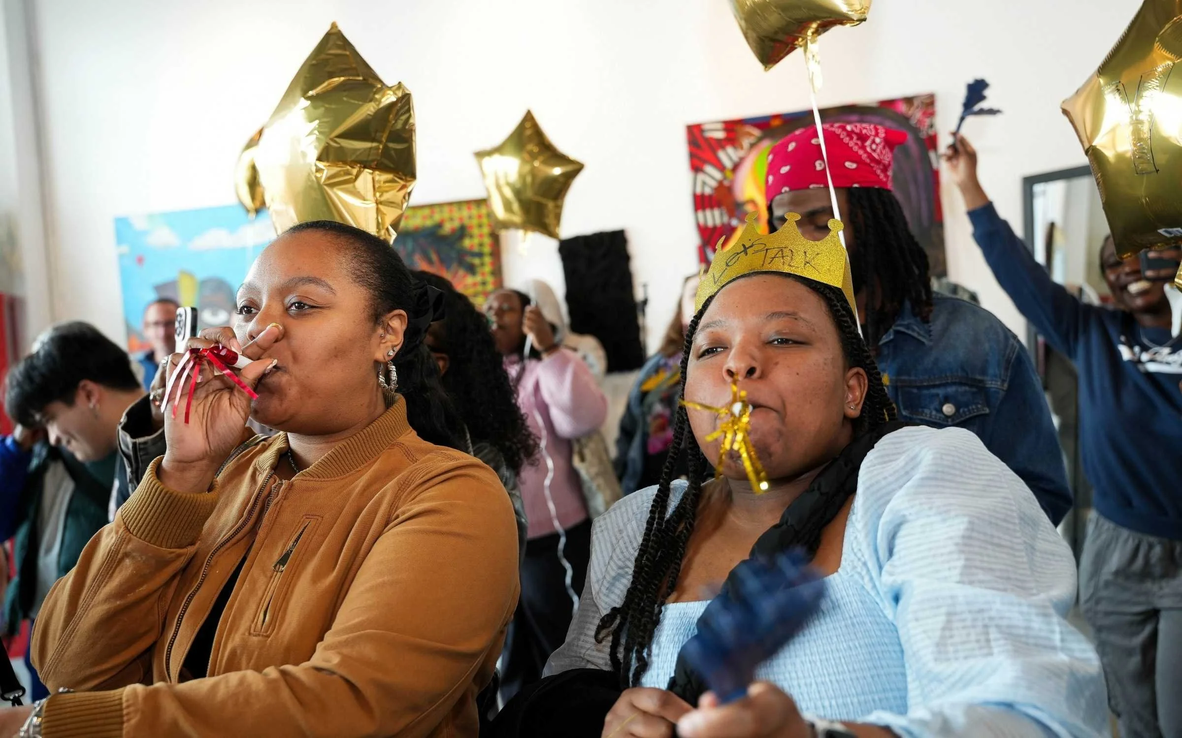 A diverse group of people celebrating indoors, wearing party hats and gold balloons, with some holding drinks and cameras.