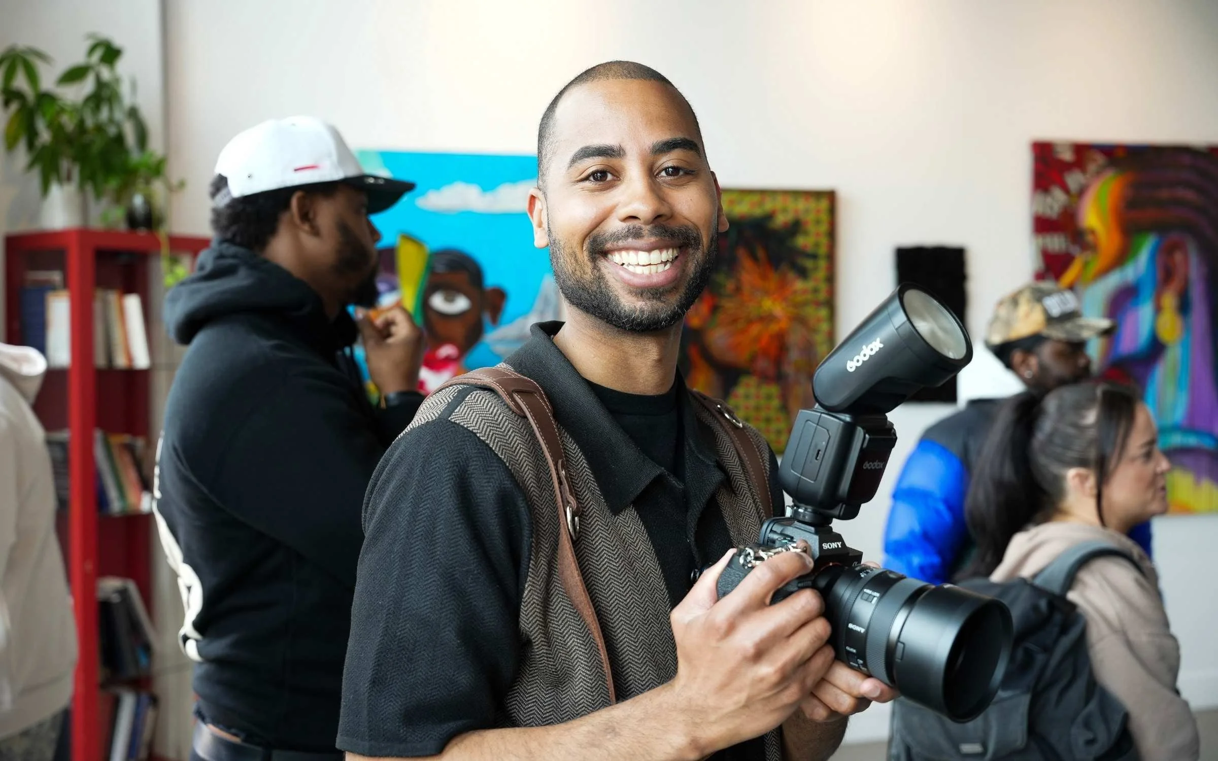 Wes Kinard smiling and holding a camera in a crowded indoor art gallery with colorful paintings and other visitors in the background.