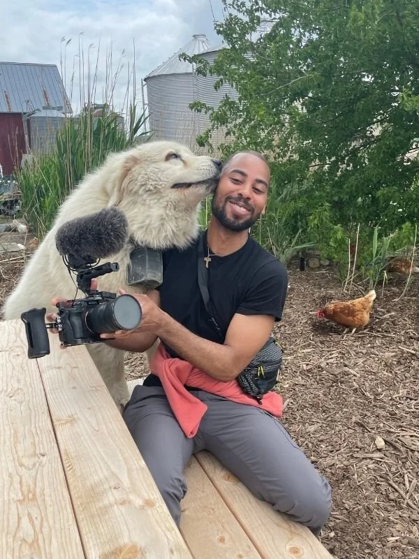 A smiling man in outdoor farm setting holding a large white dog with a camera in hand, while a chicken is visible in the background near green plants and metal silos.
