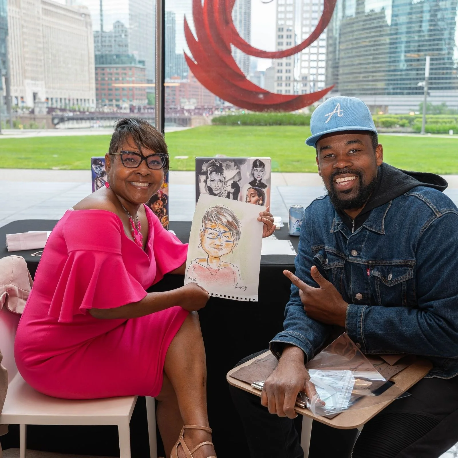 A woman in a bright pink dress and glasses holding a caricature portrait of herself, sitting next to a man in a denim jacket and a blue baseball cap, both smiling at the camera inside a building with a cityscape view in the background.