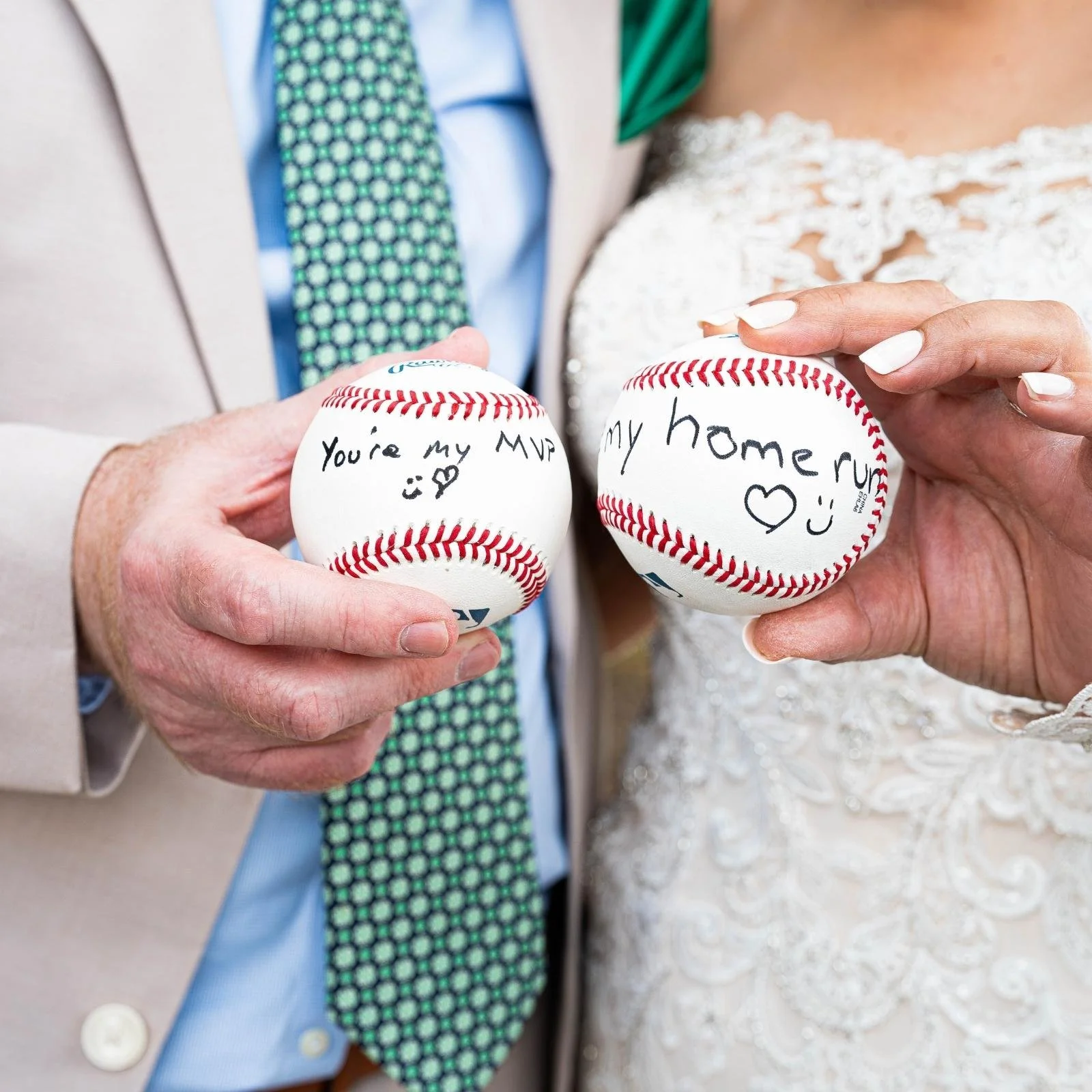Close-up of two hands holding baseballs with handwritten messages, one reads 'You're my MVP' with a smiley face, and the other reads 'my homerun' with a heart and smiley face, dressed in wedding attire.