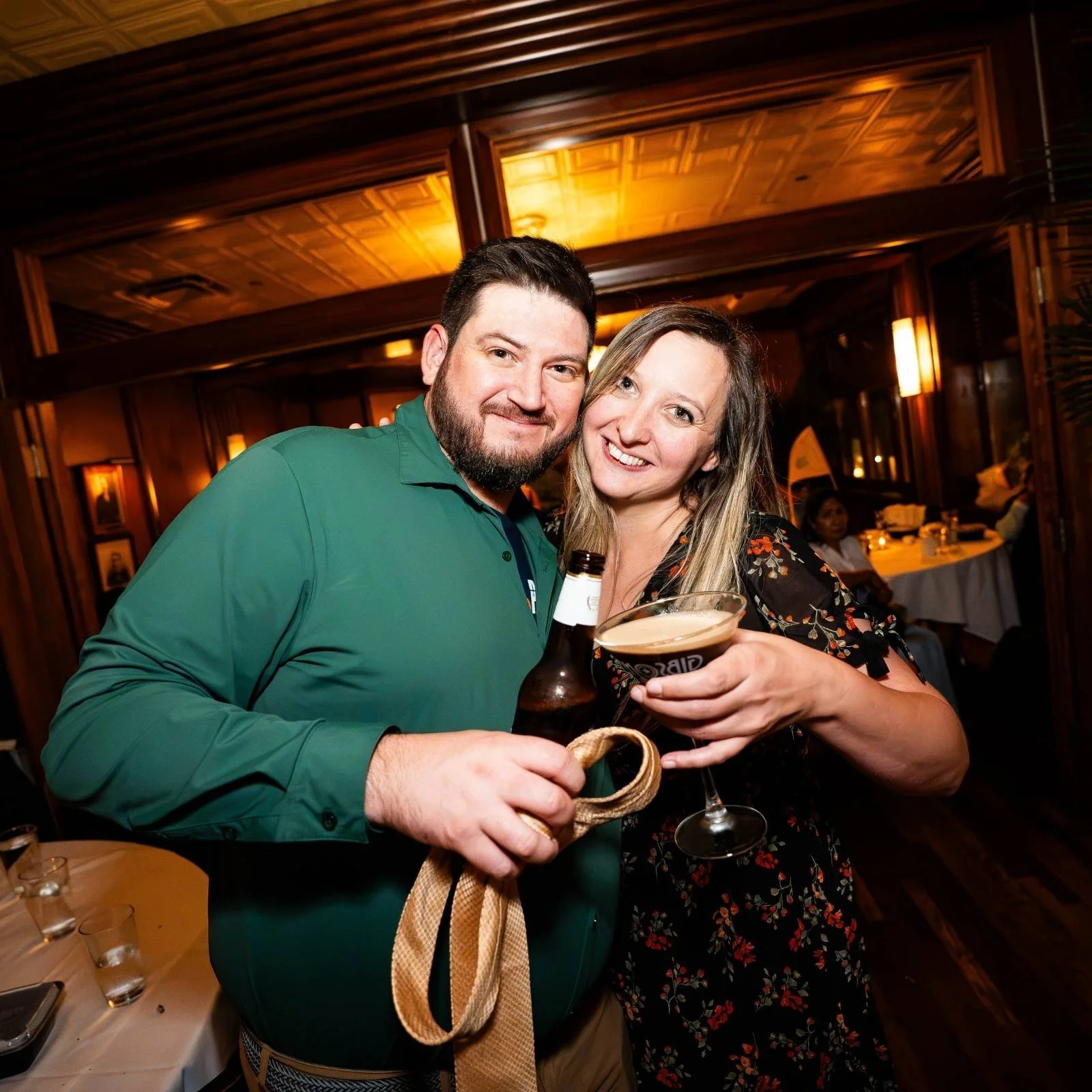 A man and woman smiling and holding drinks at a social gathering in a warmly lit, wood-paneled restaurant or bar. The woman is holding a glass of dark beer and the man has a beer bottle, with the woman also holding a purse.