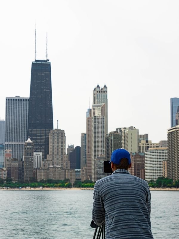 Wes Kinard wearing a blue cap and striped shirt filming or photographing the Chicago skyline with a camera, across a body of water.