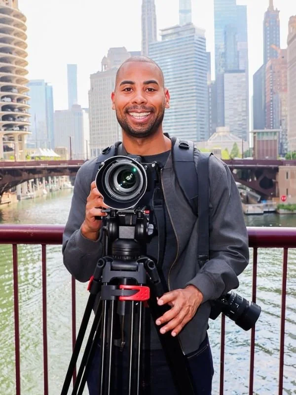 Wes Kinard holding a camera on a tripod, standing outdoors near a river with an urban Chicago skyline in the background.