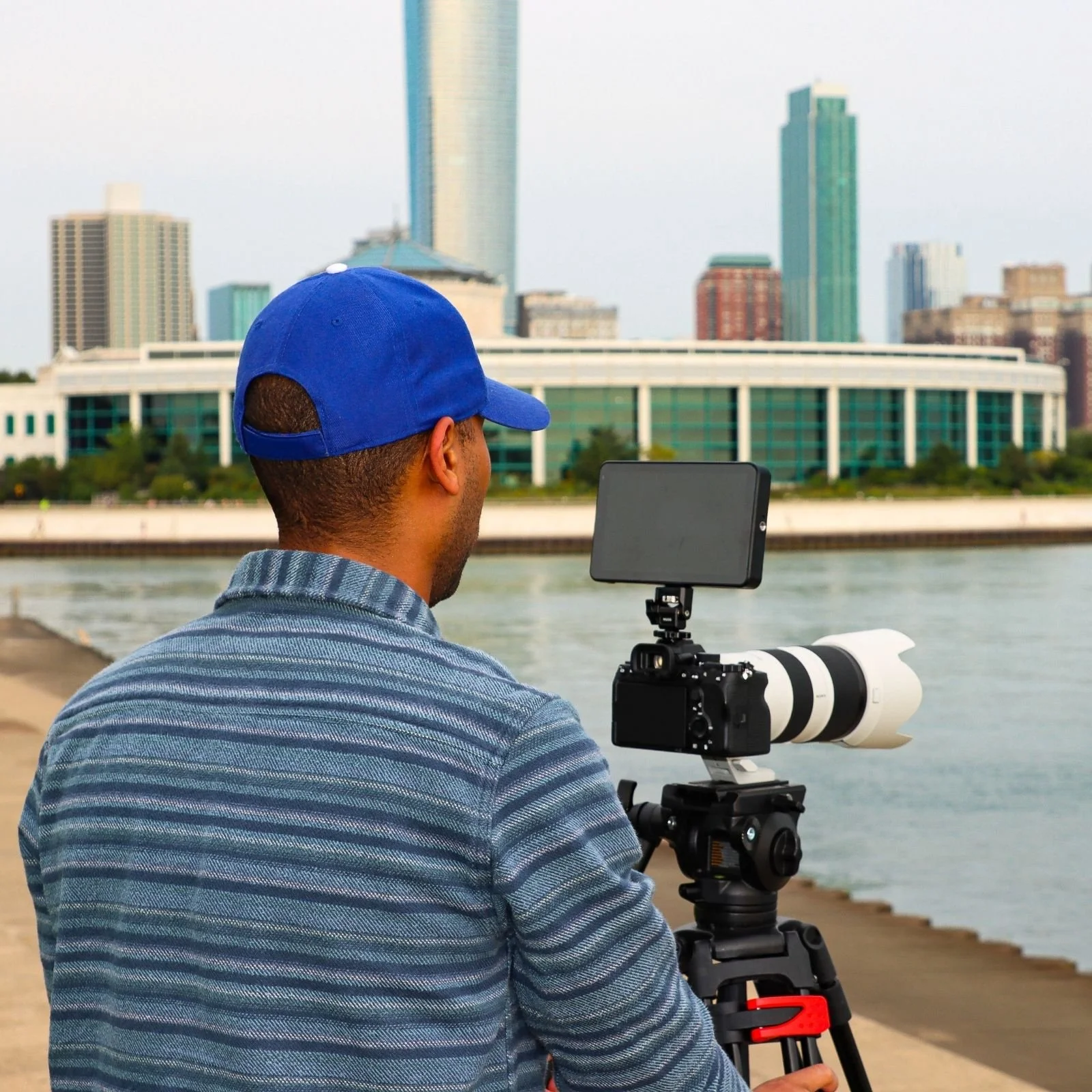 Wes Kinard wearing a blue cap and striped shirt using a camera with a telephoto lens and external monitor, by a Chicago river with city skyscrapers in background.