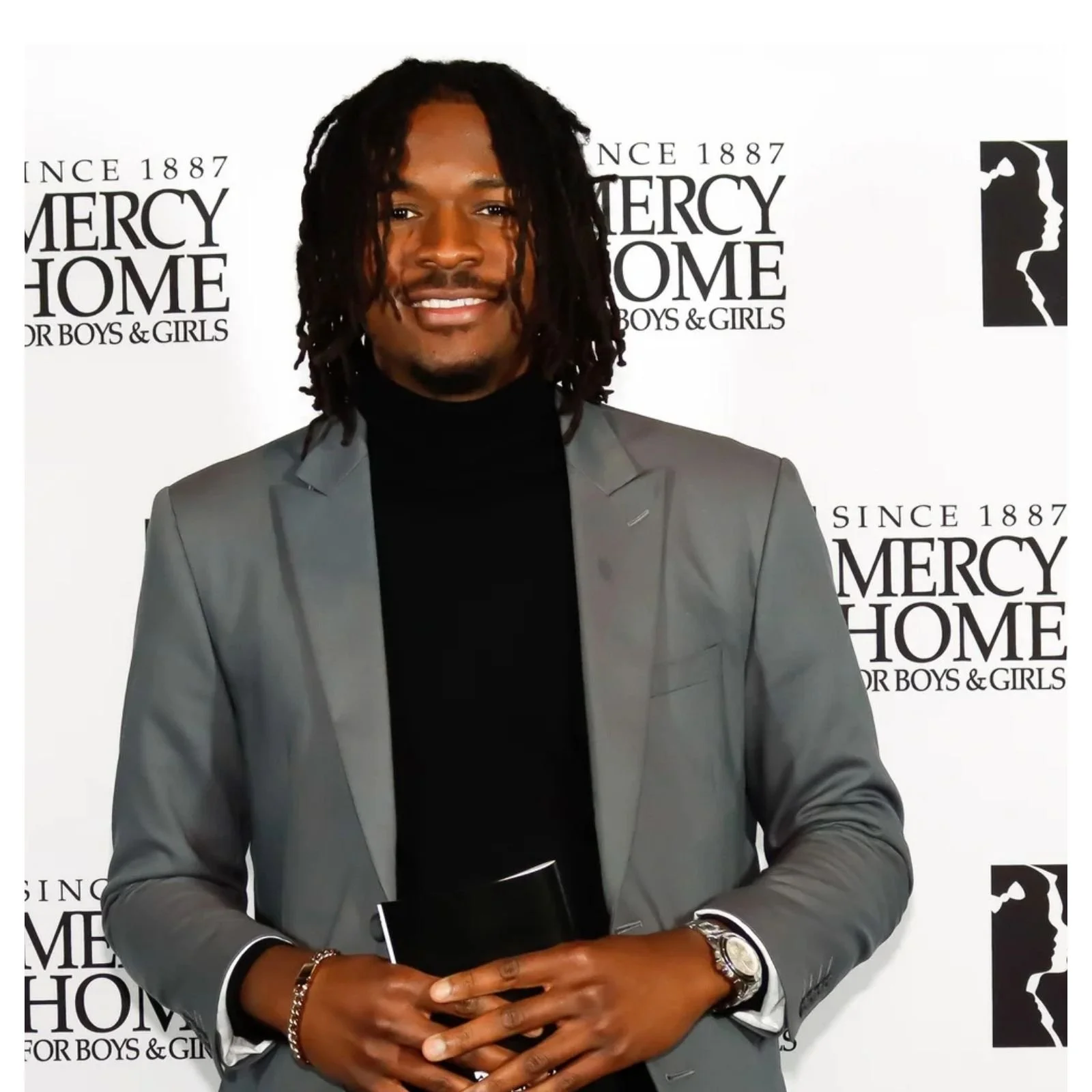 Man with dreadlocks wearing a gray suit and black turtleneck standing in front of Mercy Home for Boys & Girls backdrop.