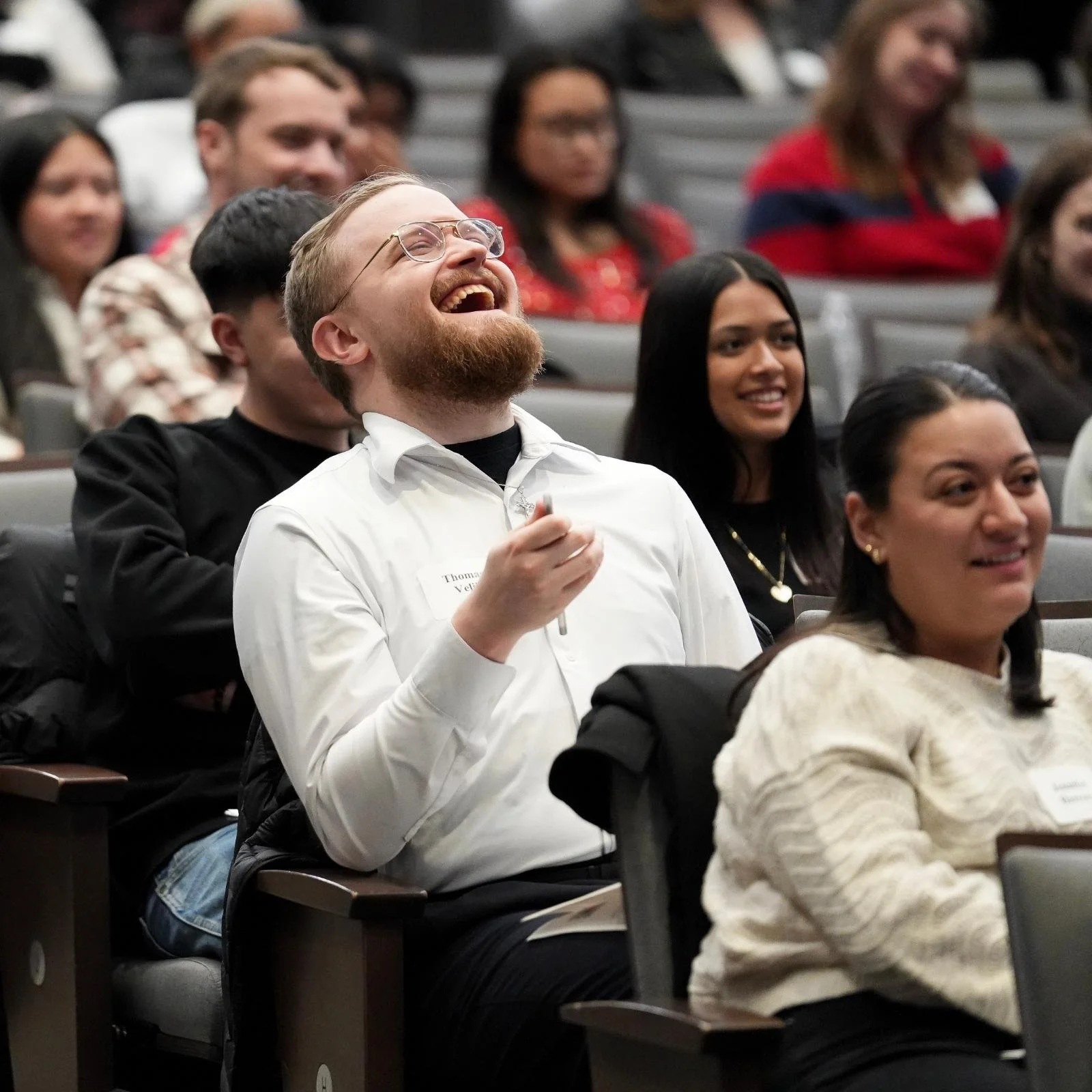People attending a conference or seminar, sitting in rowed seats, with one man in the foreground laughing and others smiling or listening, in a large indoor auditorium.