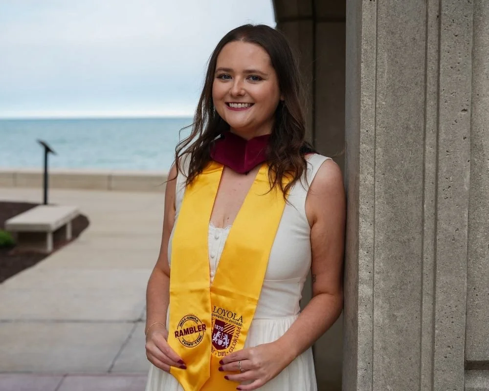 A young woman in a white dress standing outdoors near a concrete structure, smiling, with a yellow graduation sash from Loyola University and a maroon stole.
