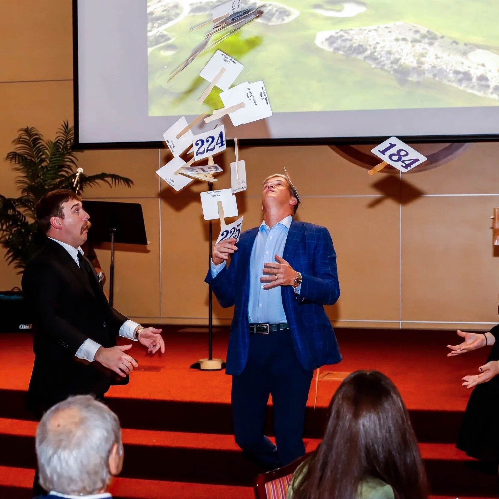 Two men are in a presentation or conference setting. One man, dressed in a black suit, is kneeling with his hands gesturing. The other man, in a blue plaid suit, is standing and looking up at the projection screen, which displays floating signs and numbers. The audience is visible in the foreground.