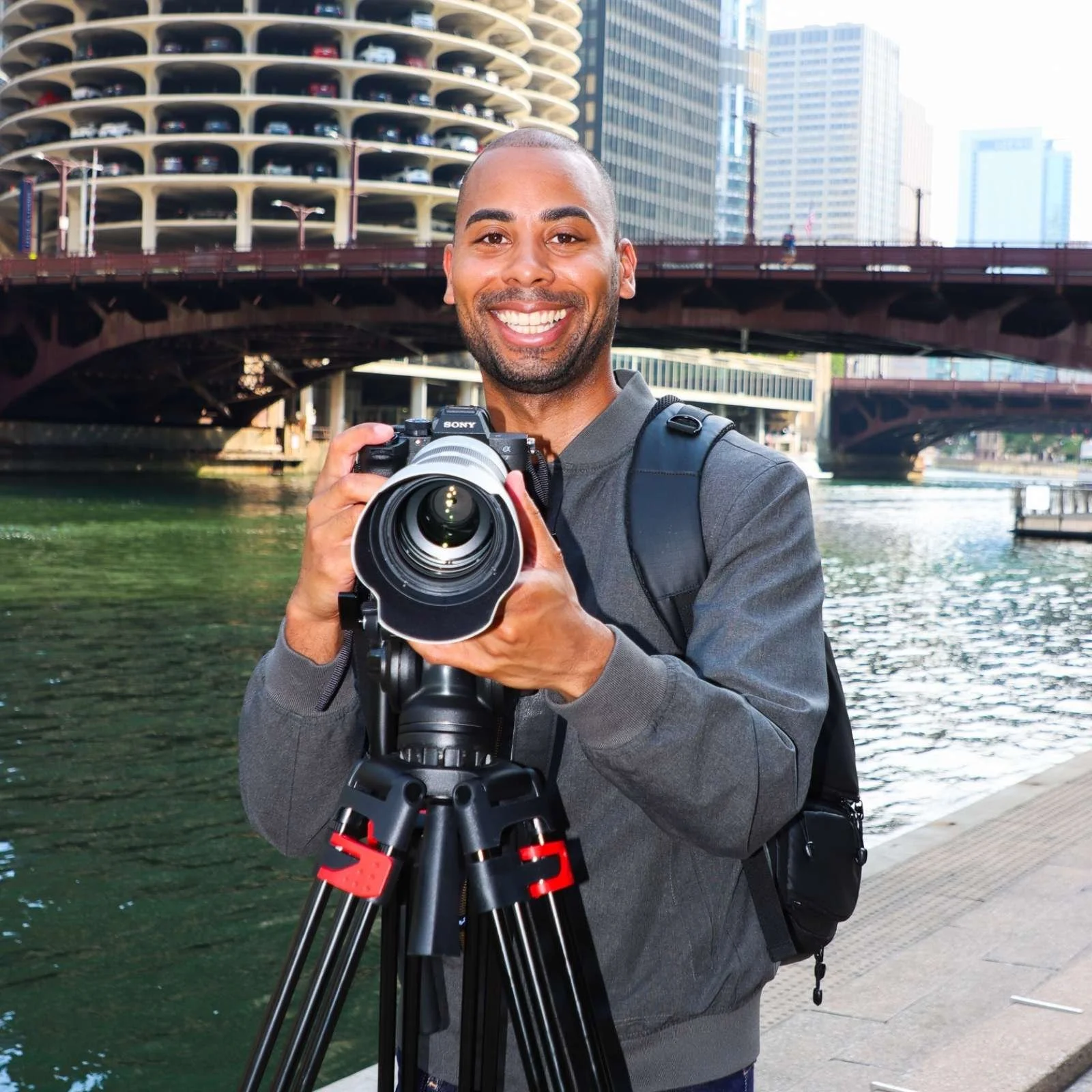 A smiling man with a beard and short hair, holding a camera on a tripod, standing next to a river in an urban area with tall buildings and a parking structure in the background.