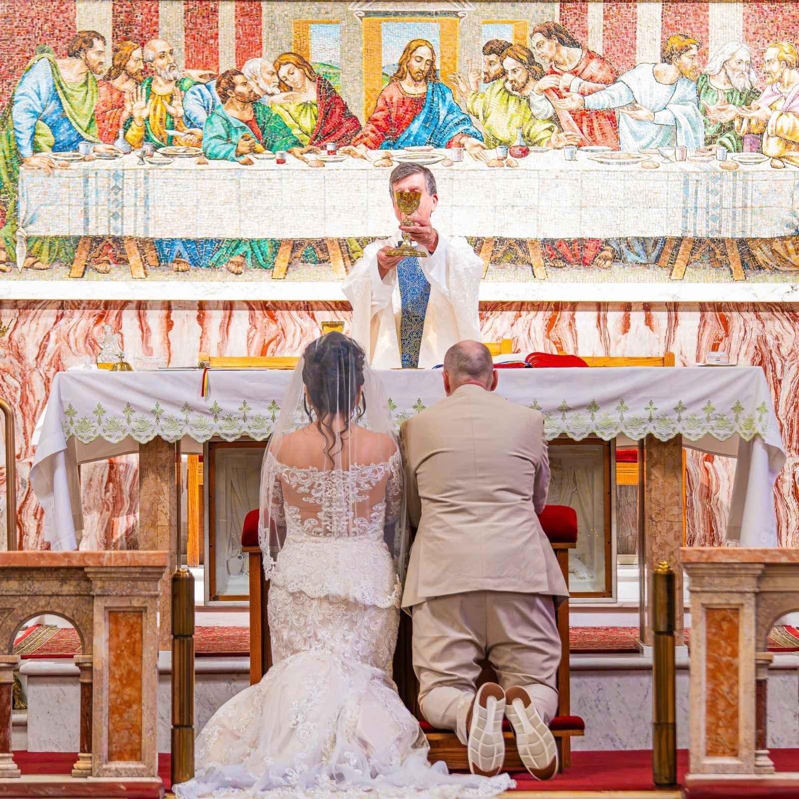 A bride and groom kneeling in front of an altar during a wedding ceremony inside a church. The bride wears a white lace wedding dress and veil, and the groom wears a light-colored suit. Both are kneeling on red cushions facing the altar, with their backs to the camera. A priest is standing in front of them holding a chalice, and a large colorful mosaic depicting Jesus at the Last Supper hangs behind them.