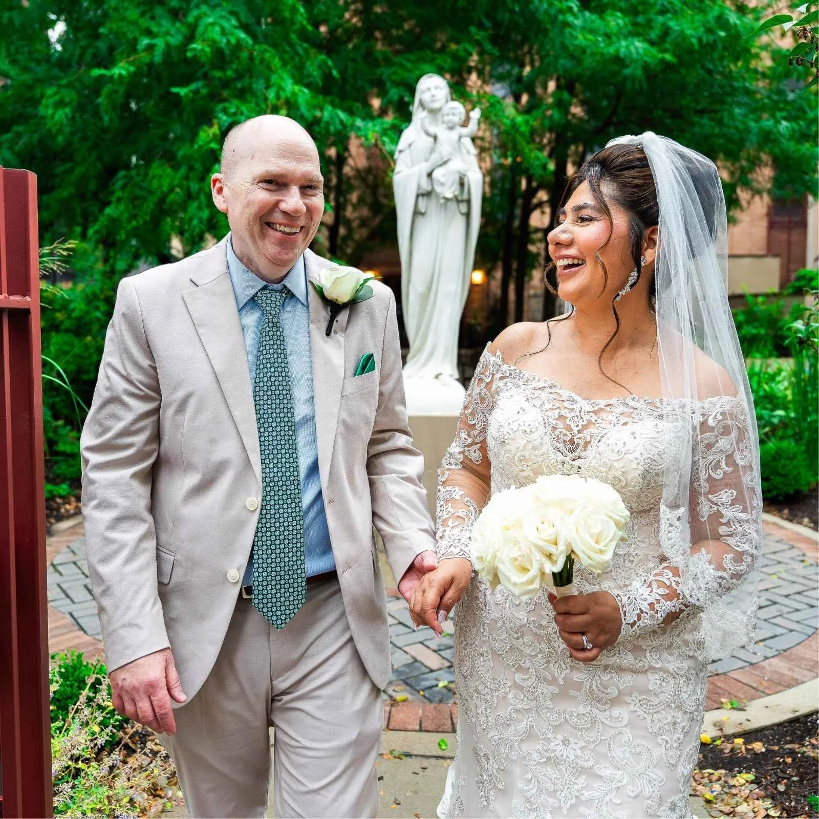 A bride and groom holding hands outdoors, smiling. The bride wears a lace wedding dress with a veil, holding a bouquet of white roses. The groom wears a beige suit with a light blue shirt, patterned tie, and a boutonniere. In the background, there is a white statue of a woman with a child, surrounded by greenery.