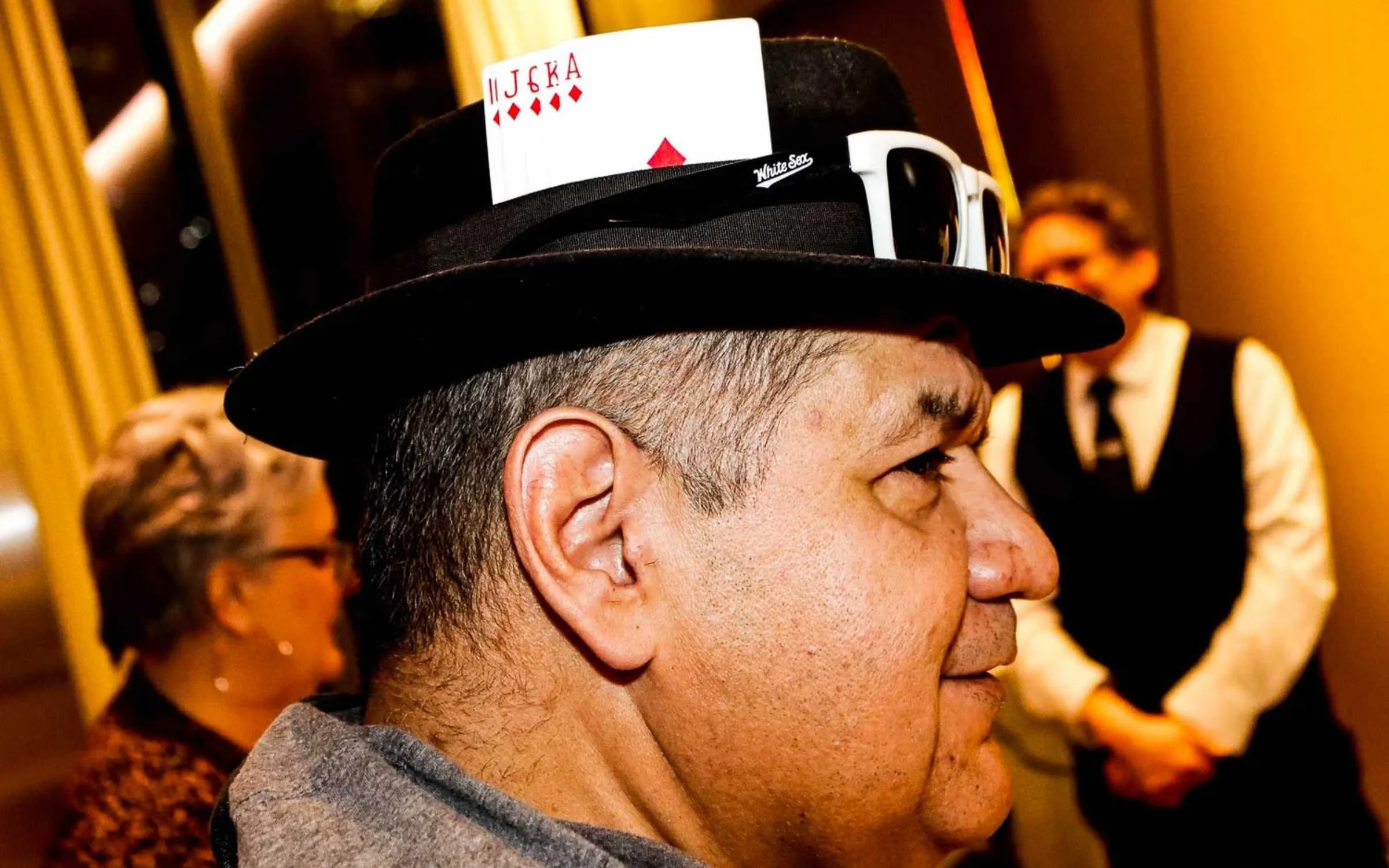 A man wearing a black hat with a card tucked into its band, and sunglasses, standing indoors with other people around him.