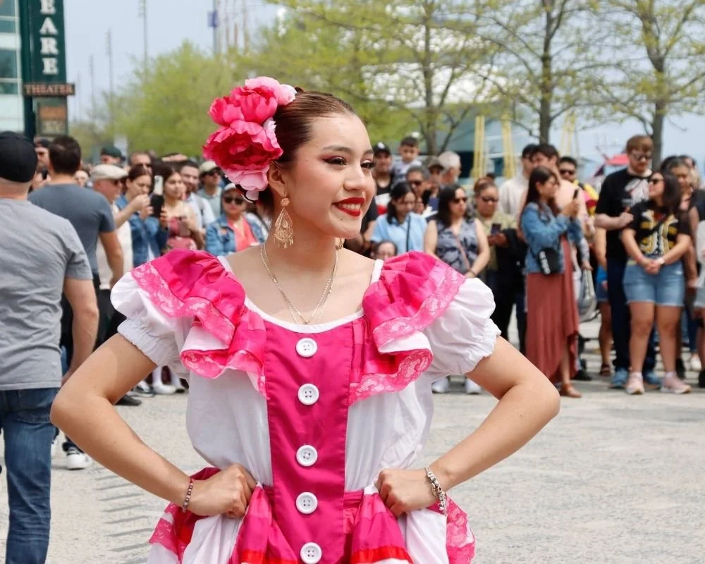Young woman dressed in traditional Mexican attire with pink flowers in her hair, standing in front of a crowd at a festival or event outdoors.
