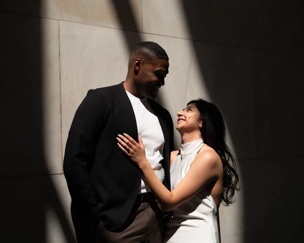 A couple smiling and looking at each other, standing close together near a wall with shadows cast on it, in a well-lit indoor setting.