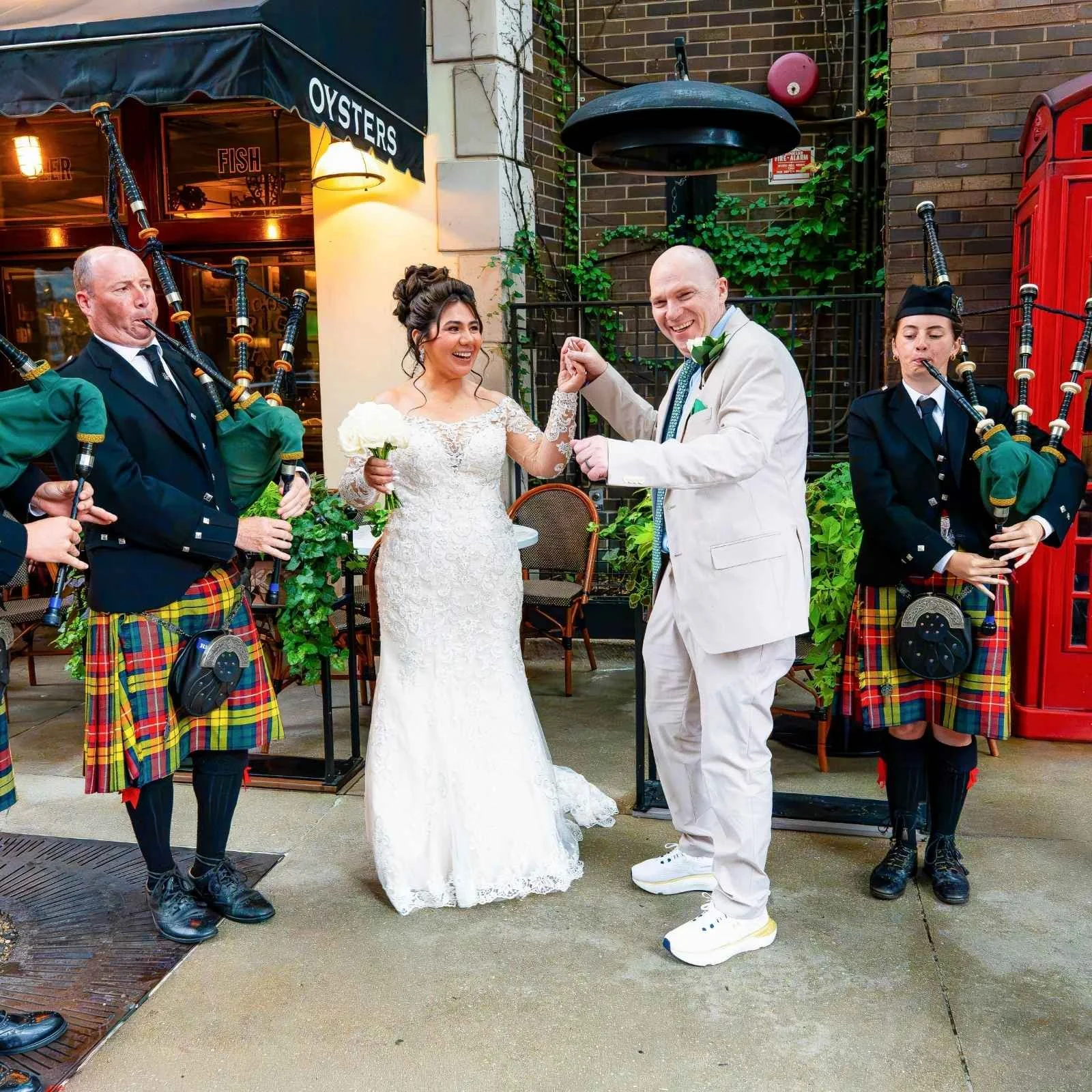A bride and groom dance together at their outdoor wedding celebration, surrounded by musicians playing bagpipes, in front of a brick building with a sign for oysters and fish.