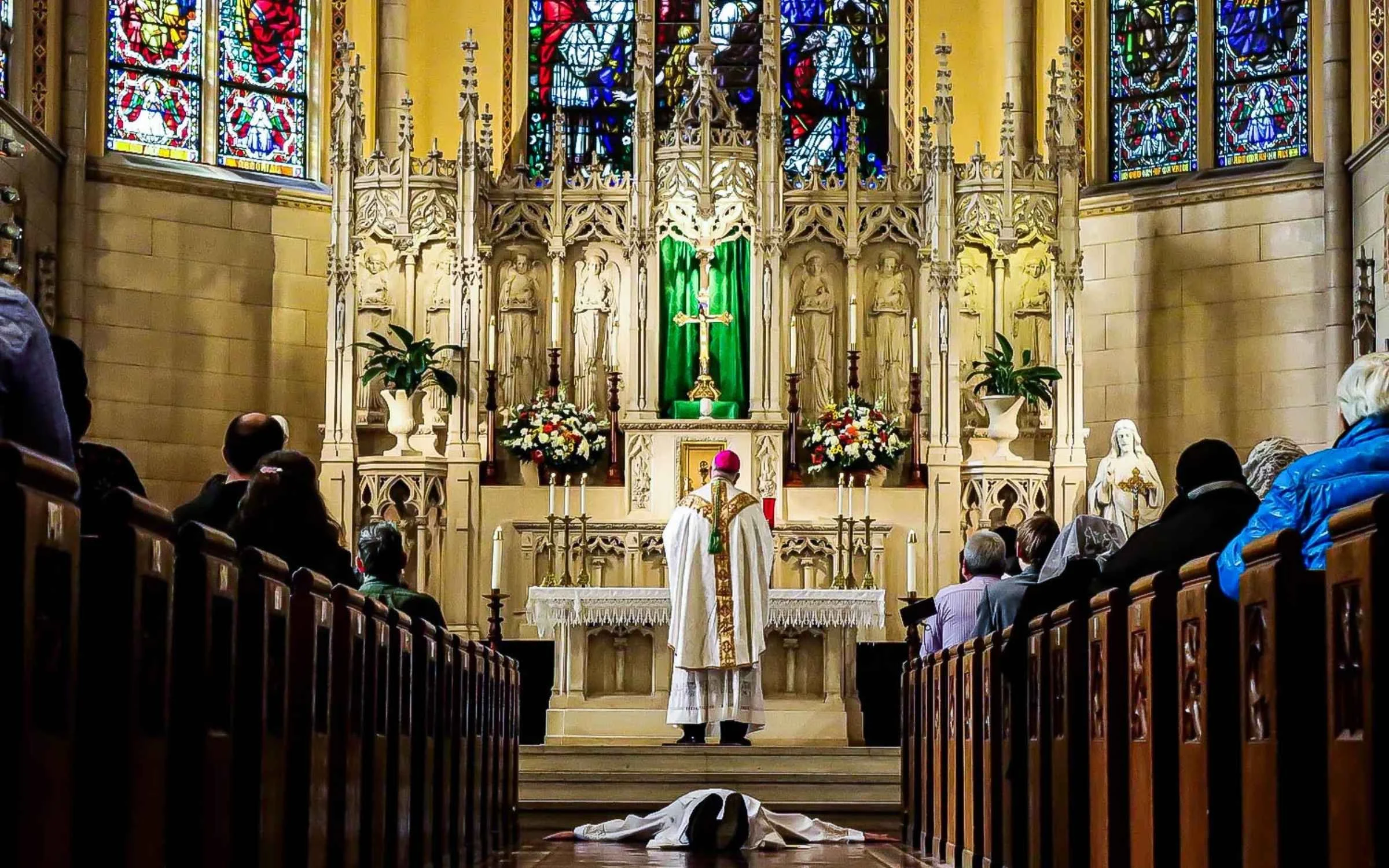 Interior of a church with stained glass windows, an altar, and a priest with a choir or congregation in the pews.