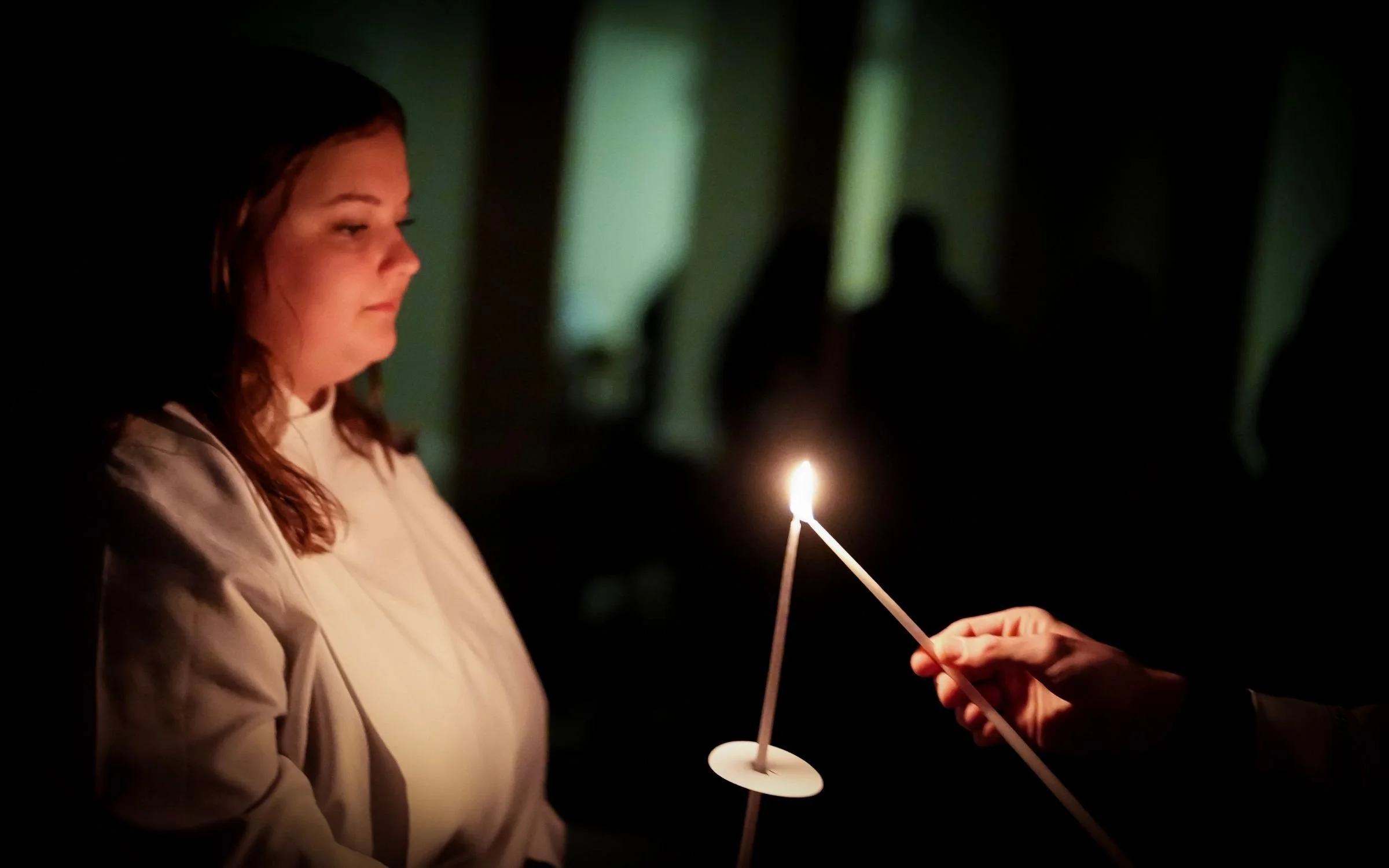 A woman with long, wavy hair and a white shirt holding a small lit candle, with another hand helping her hold it, against a dark background.