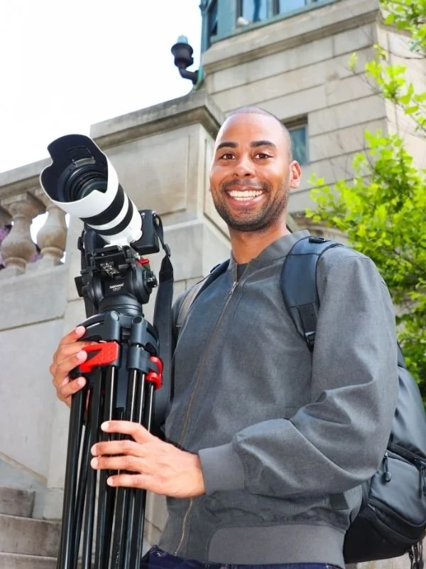 Wes Kinard holding a professional camera with a tripod and a telephoto lens, standing outdoors in front of a stone building and green trees.