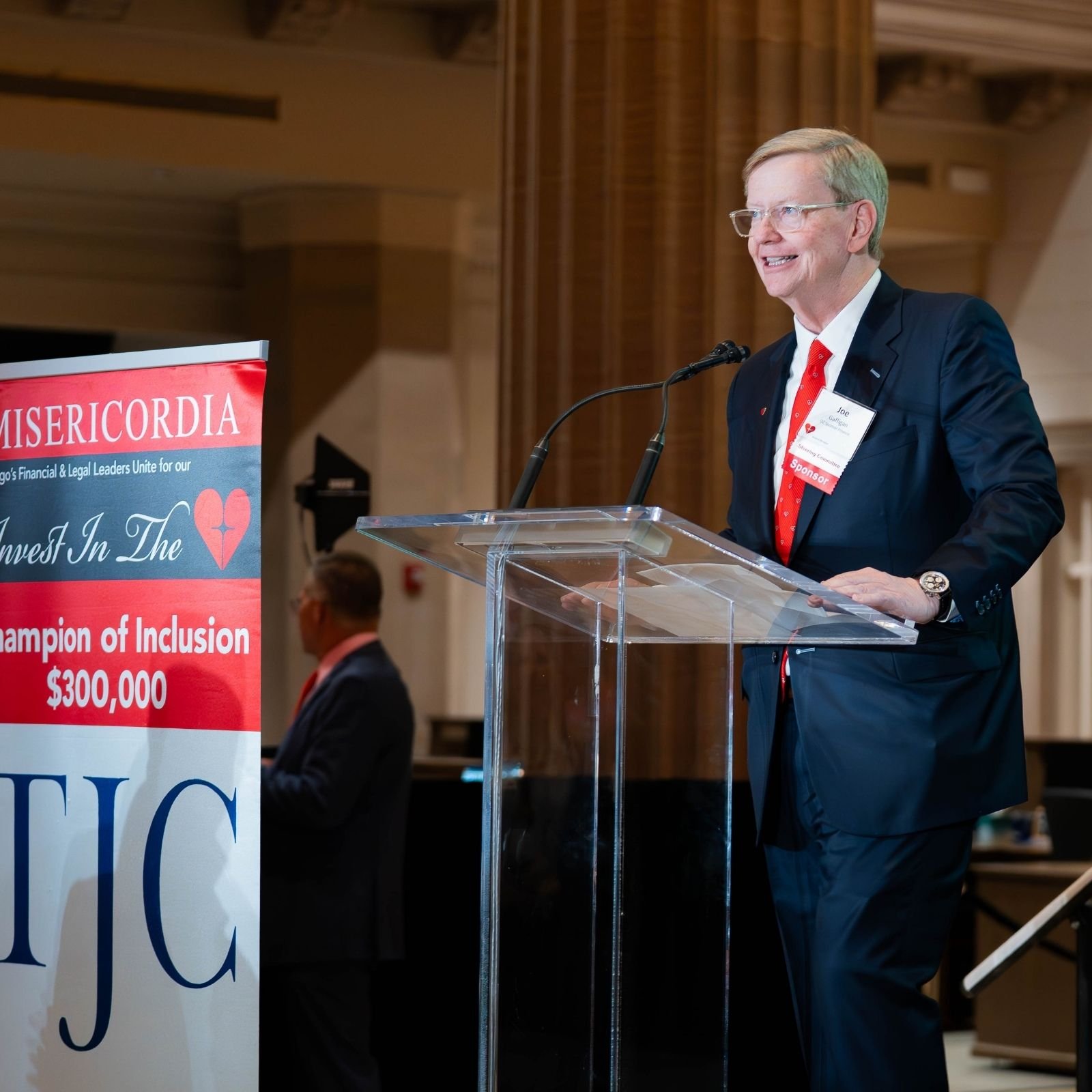 A man in a suit with glasses and a red tie speaking at a podium with a microphone, at an event with a large red and blue banner that reads 'MISERICORDIA' and mentions a donation amount of $300,000.