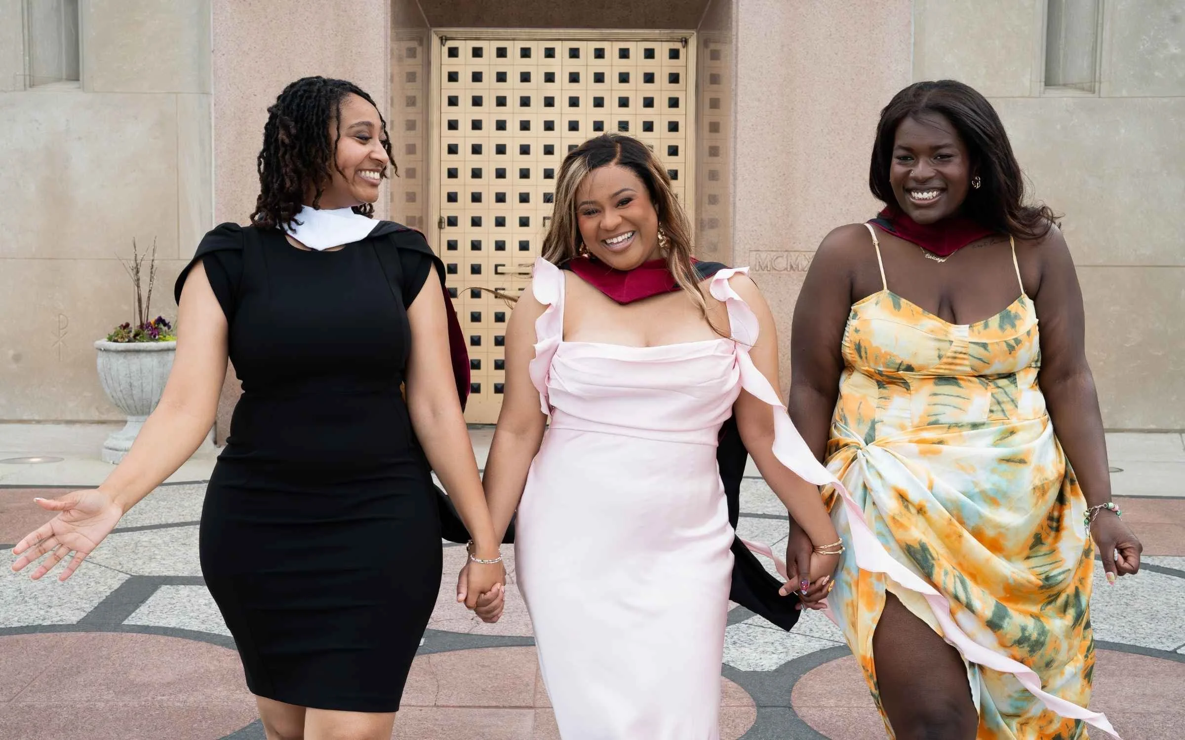 Three women holding hands and smiling outside a building with a decorative wall and planters