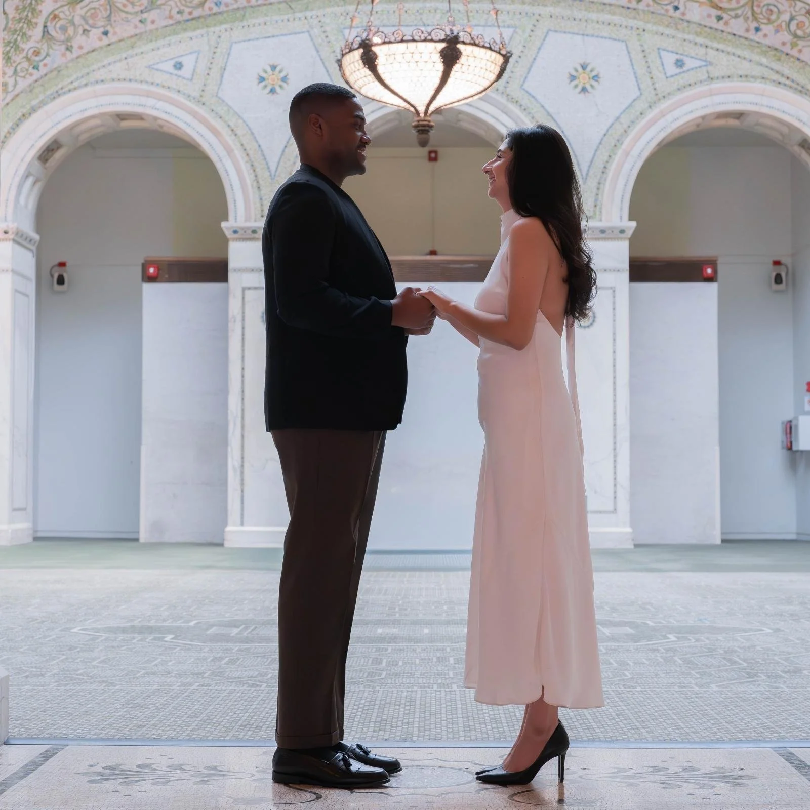 A man and woman are holding hands and facing each other in a hallway with ornate arches and a chandelier overhead. The man is wearing a black suit and brown pants, the woman is wearing a pale pink sleeveless dress and black heels.