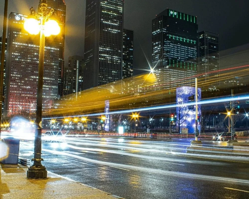 Nighttime city street with tall illuminated skyscrapers, streaks of light from moving vehicles, and vintage style street lamps.