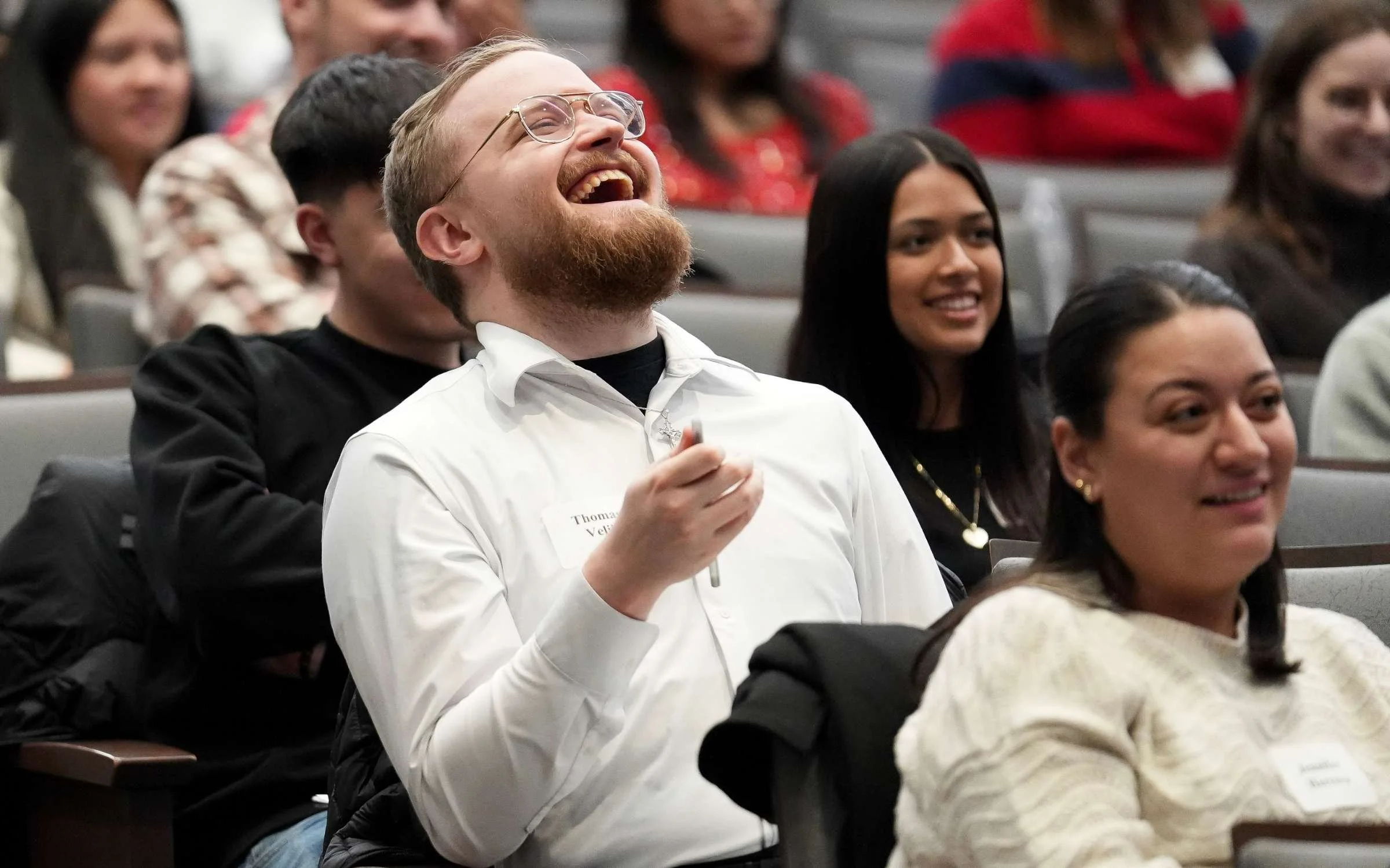A man with glasses, a beard, and a white shirt laughing and holding a pen in a conference room filled with seated people