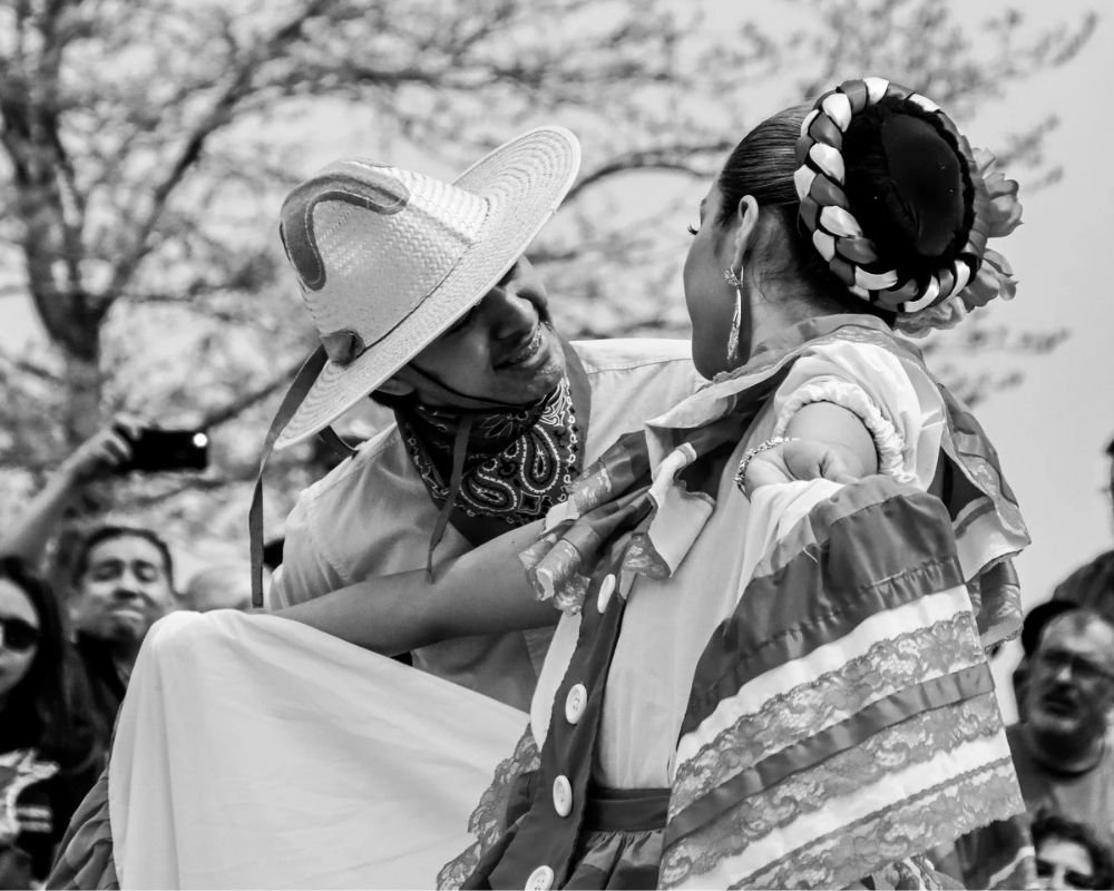 A woman wearing traditional Mexican clothing, including a large floral headpiece, and a man wearing a mexicano hat and bandana, dance together outdoors with onlookers in the background.