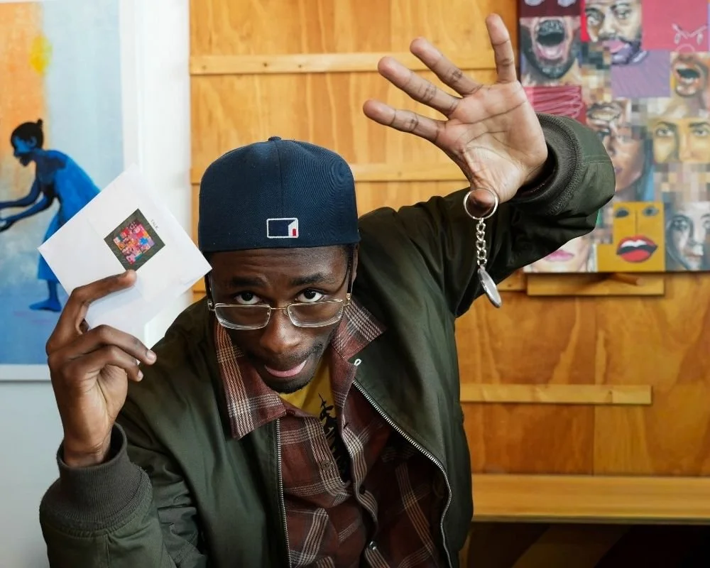 A man wearing glasses and a baseball cap holding a small paper with digital art, raising his other hand with fingers spread in a playful gesture.