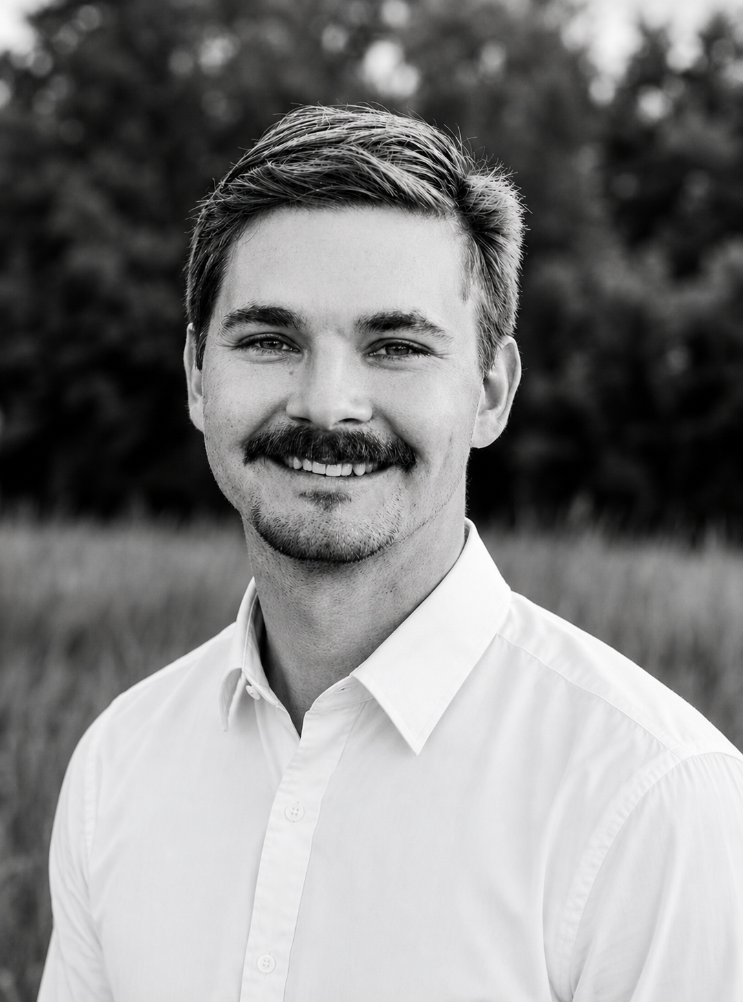 Black and white portrait of a young man with hair combed to the side, a beard, and mustache, smiling, wearing a white button-up shirt, outdoors with trees and grass in the background.