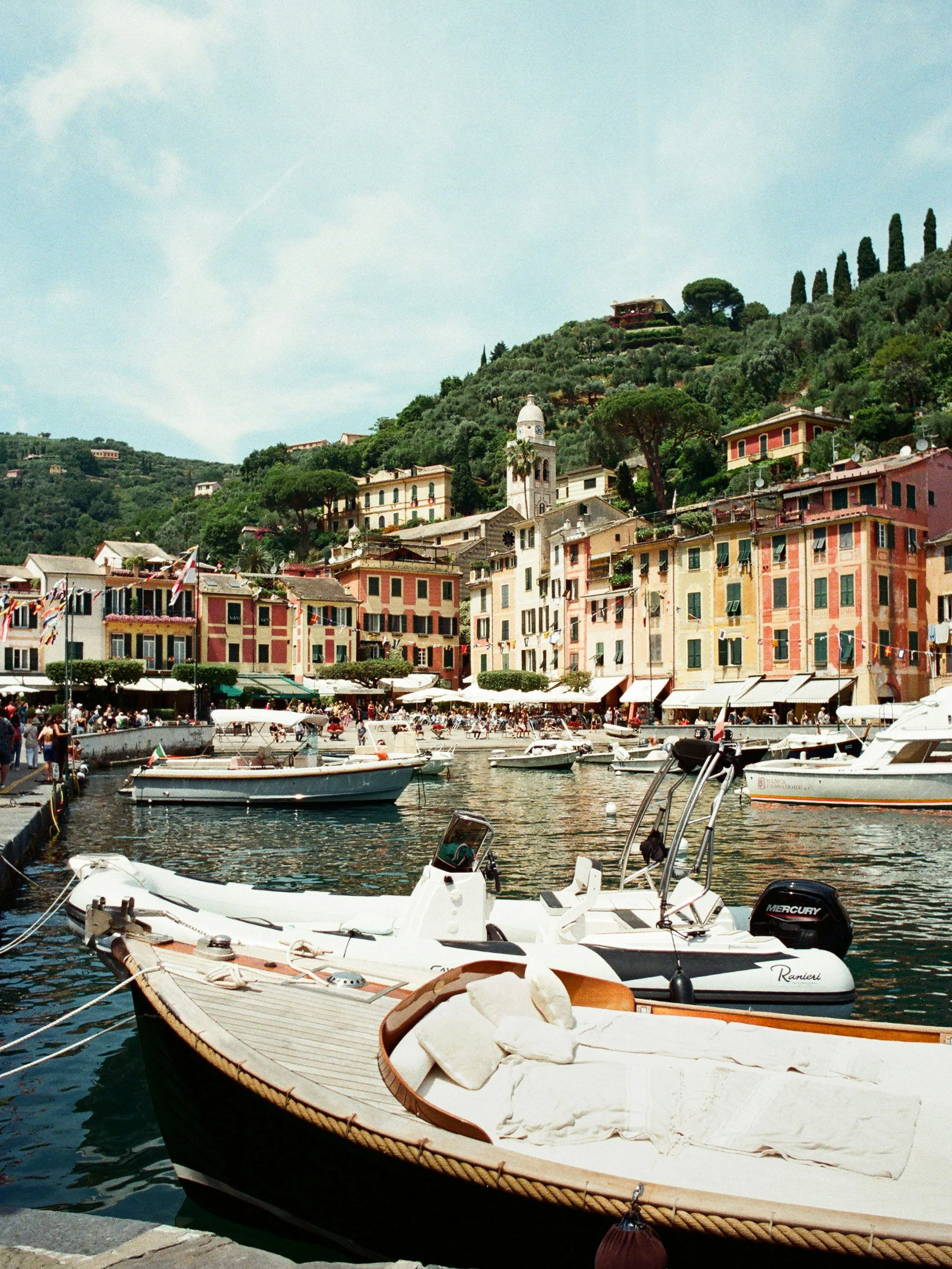 Boats docked at a harbor with colorful buildings and hillside in the background.