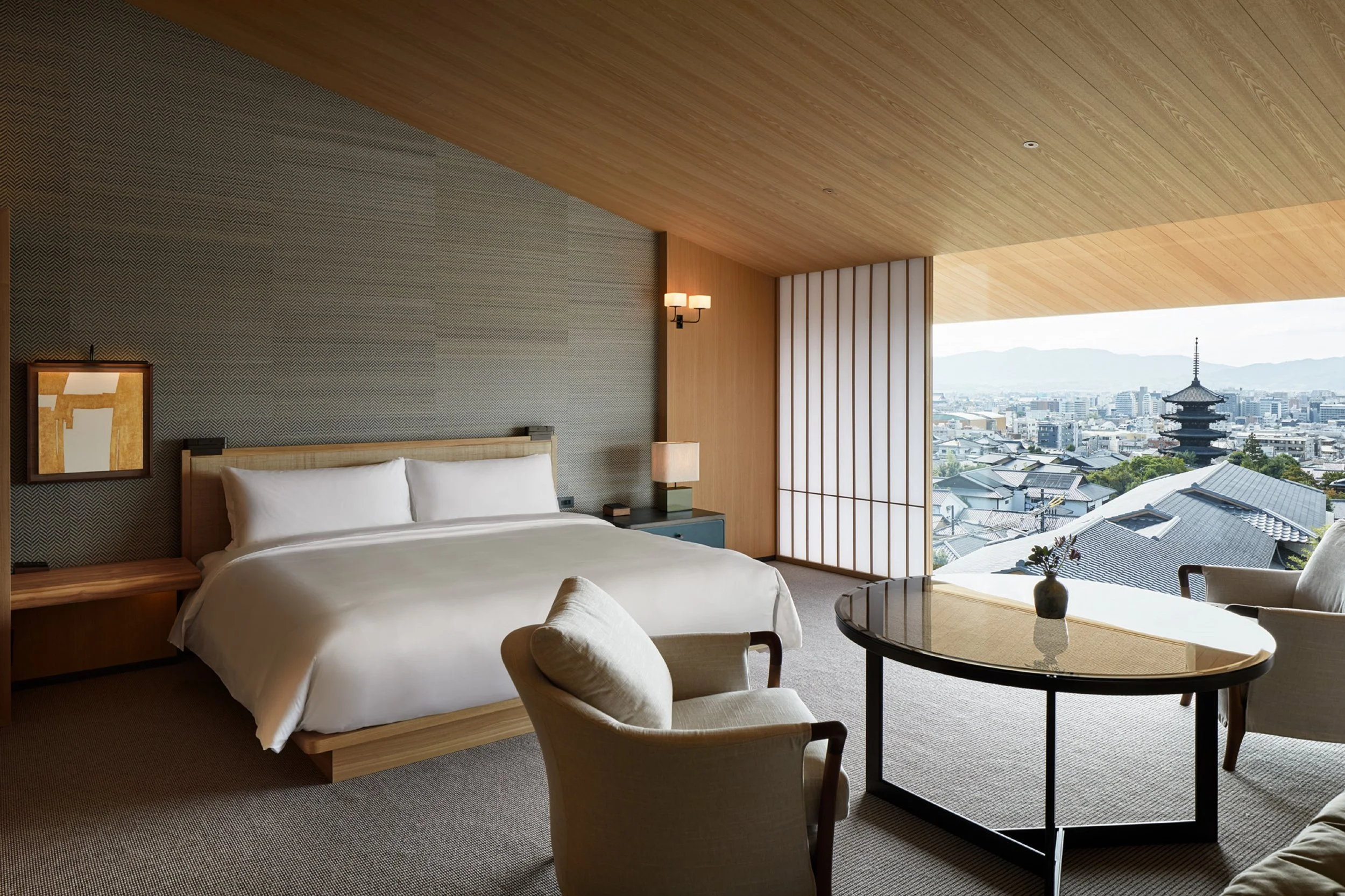 A modern hotel room with a bed, a side table, and a lounge chair, featuring large windows with a view of a cityscape and a traditional pagoda-style building in the distance.