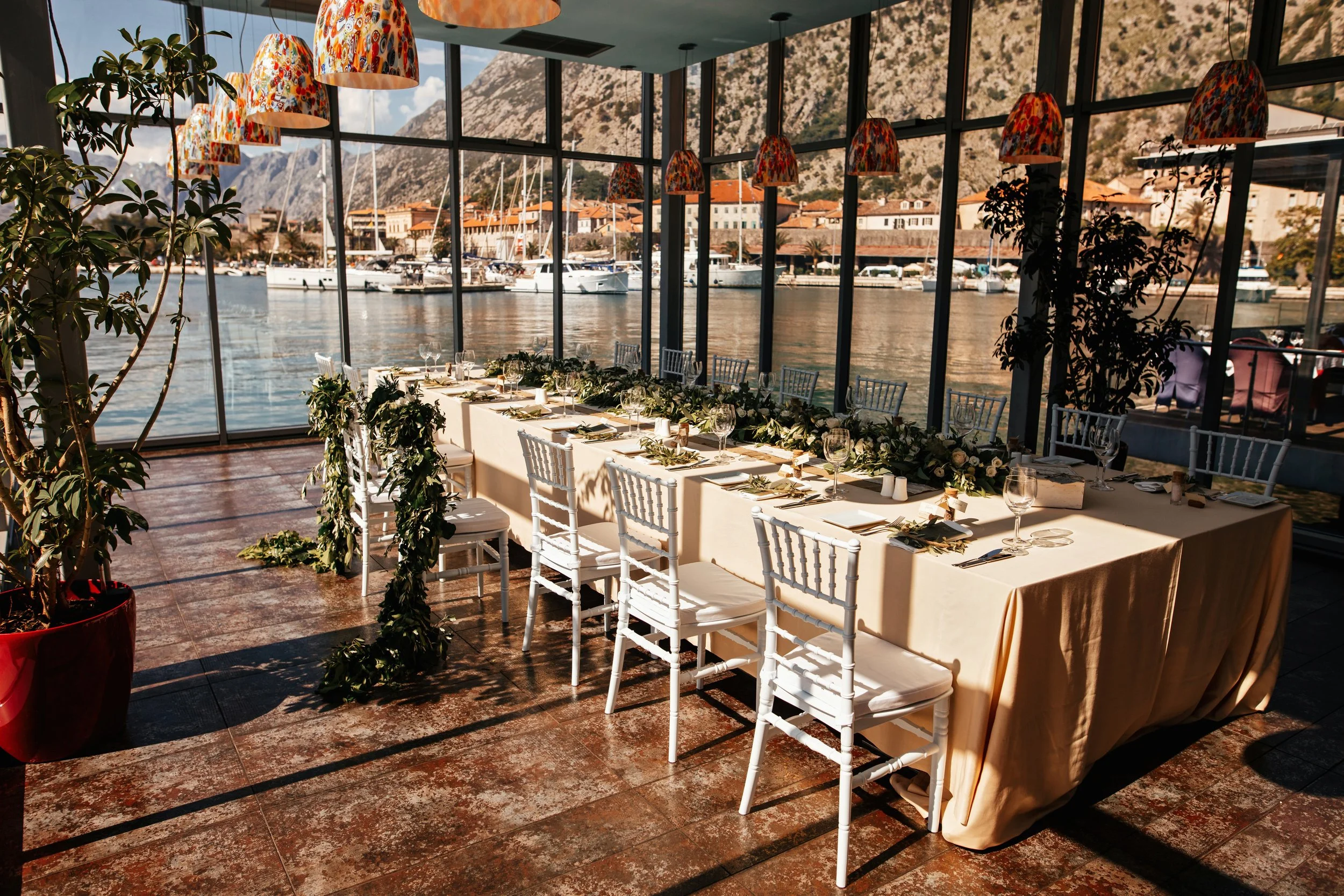 A dining table set for a group event with white chairs, wine glasses, and a green leafy garland, inside a glass-walled venue overlooking a marina with boats and a mountain backdrop.