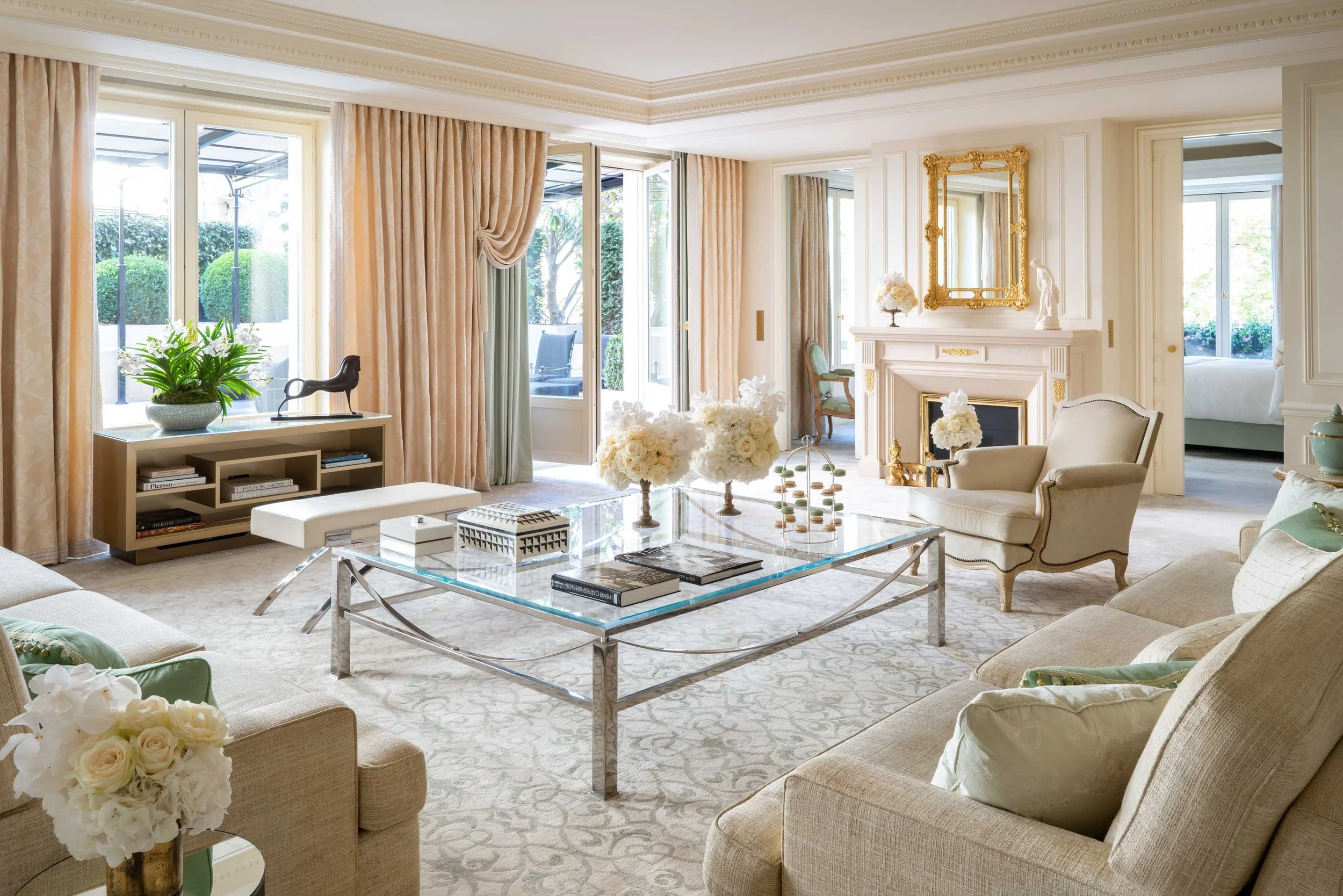 Elegant hotel suite with cream-colored sofas, a glass coffee table, large windows with beige curtains, a gold-framed mirror above a white fireplace, floral arrangements, and decorative statues.