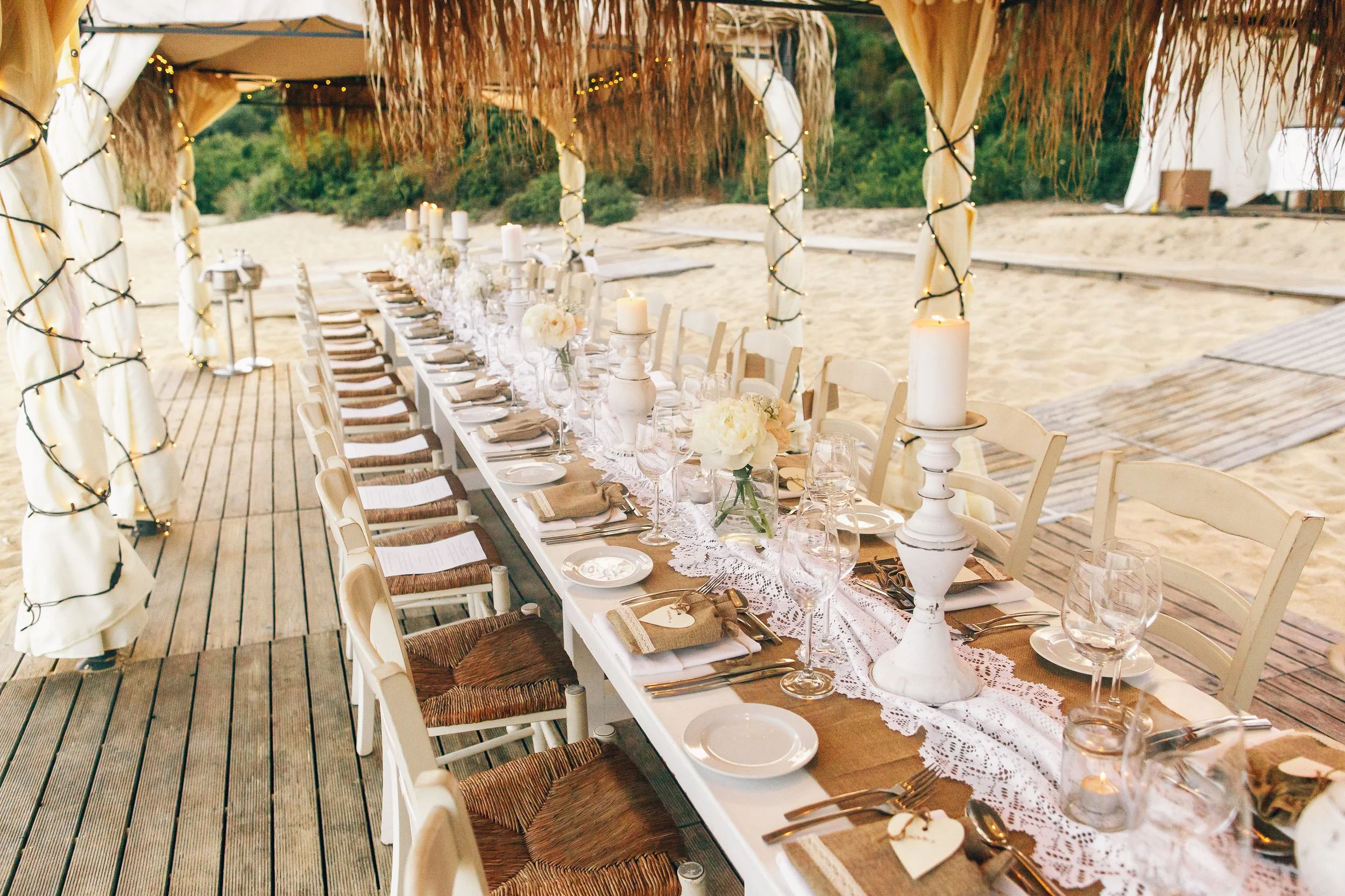 A long dining table set for a special occasion on a wooden deck with a beach background. The table is decorated with white flowers, candles, and elegant tableware, under a canopy with fabric and string lights.